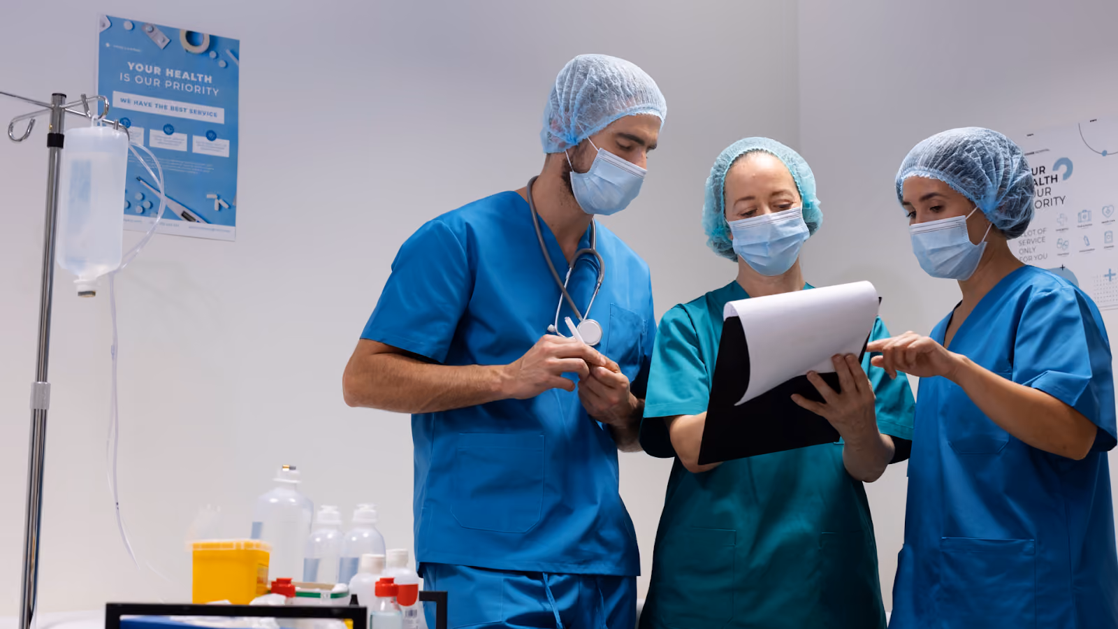 Three medical professionals in scrubs are looking at a document on a clipboard in a hospital setting with an IV stand nearby.