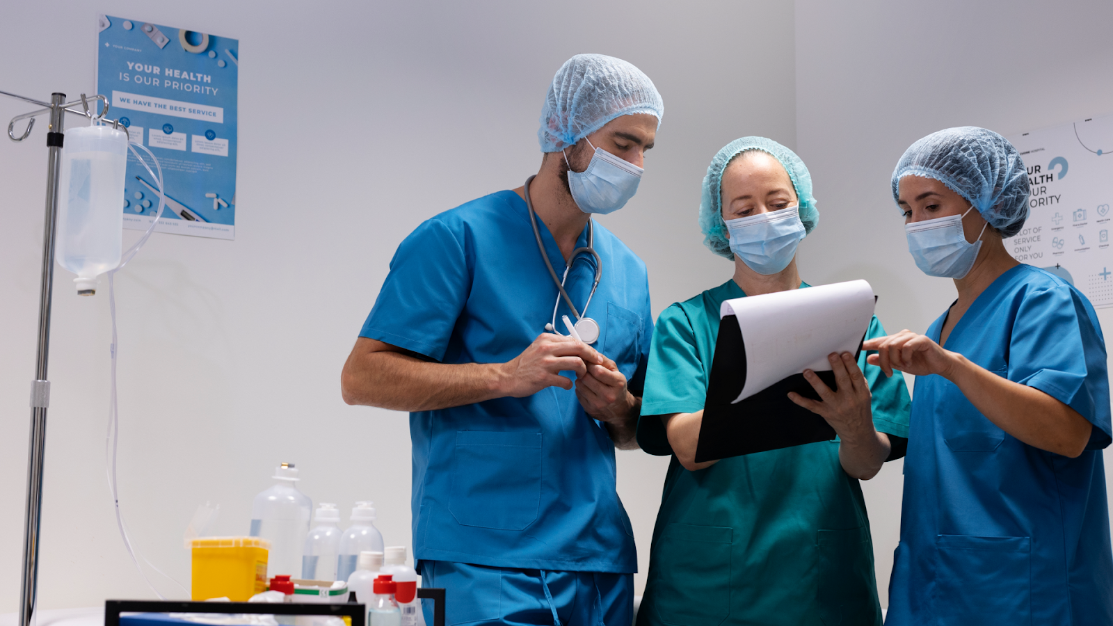 Three medical professionals in scrubs are looking at a document on a clipboard in a hospital setting with an IV stand nearby.