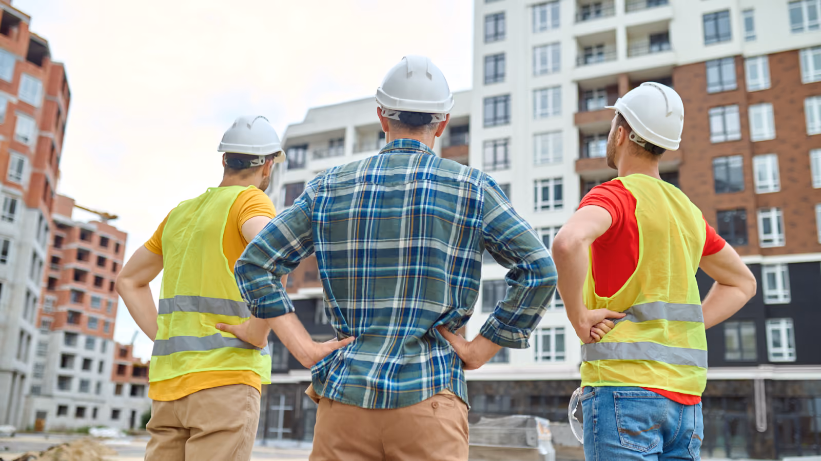 Three construction workers in hard hats and safety vests look up at a multi-story building under development.