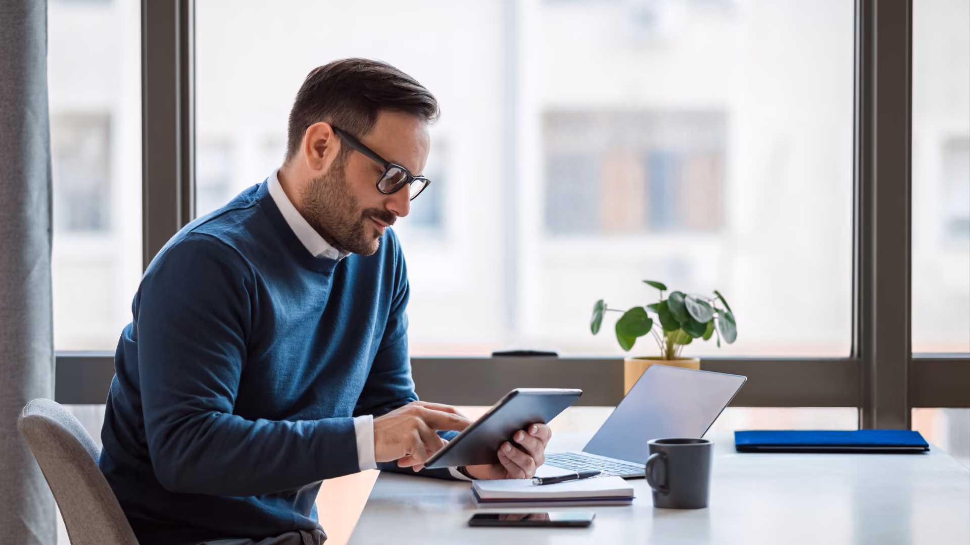 A man in a blue sweater using a tablet for digital workforce management reporting by a window.