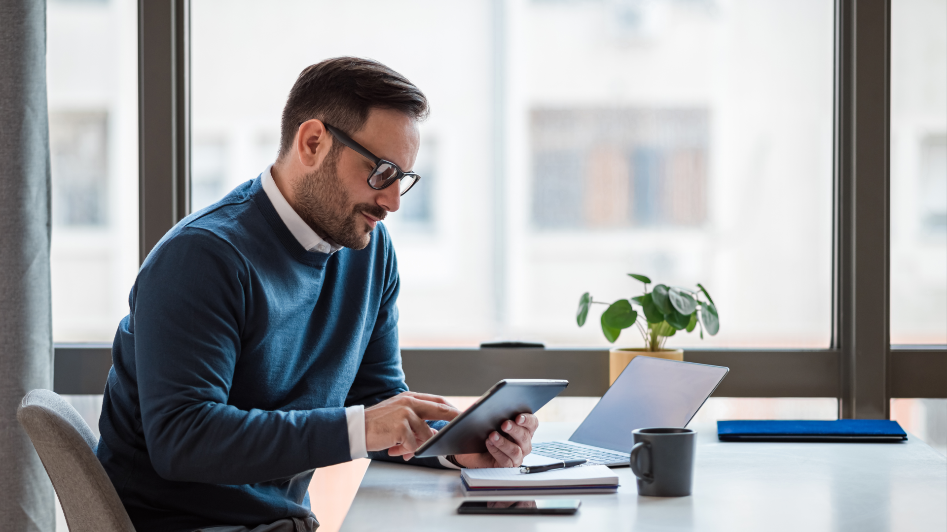 A man in a blue sweater using a tablet for digital workforce management reporting by a window.