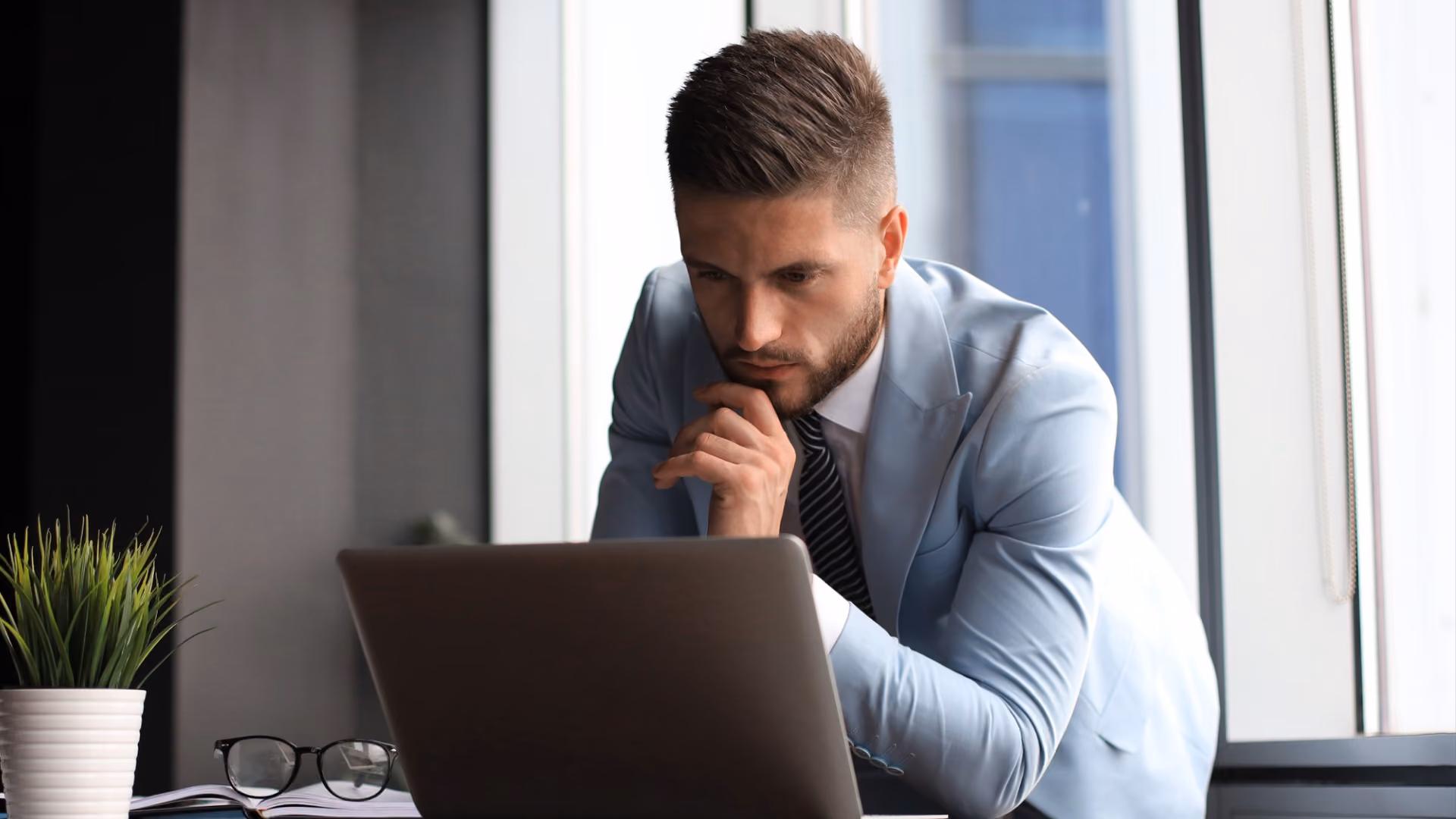 A businessman in a blue suit thoughtfully compares workforce management vs workforce optimization while looking at his laptop.