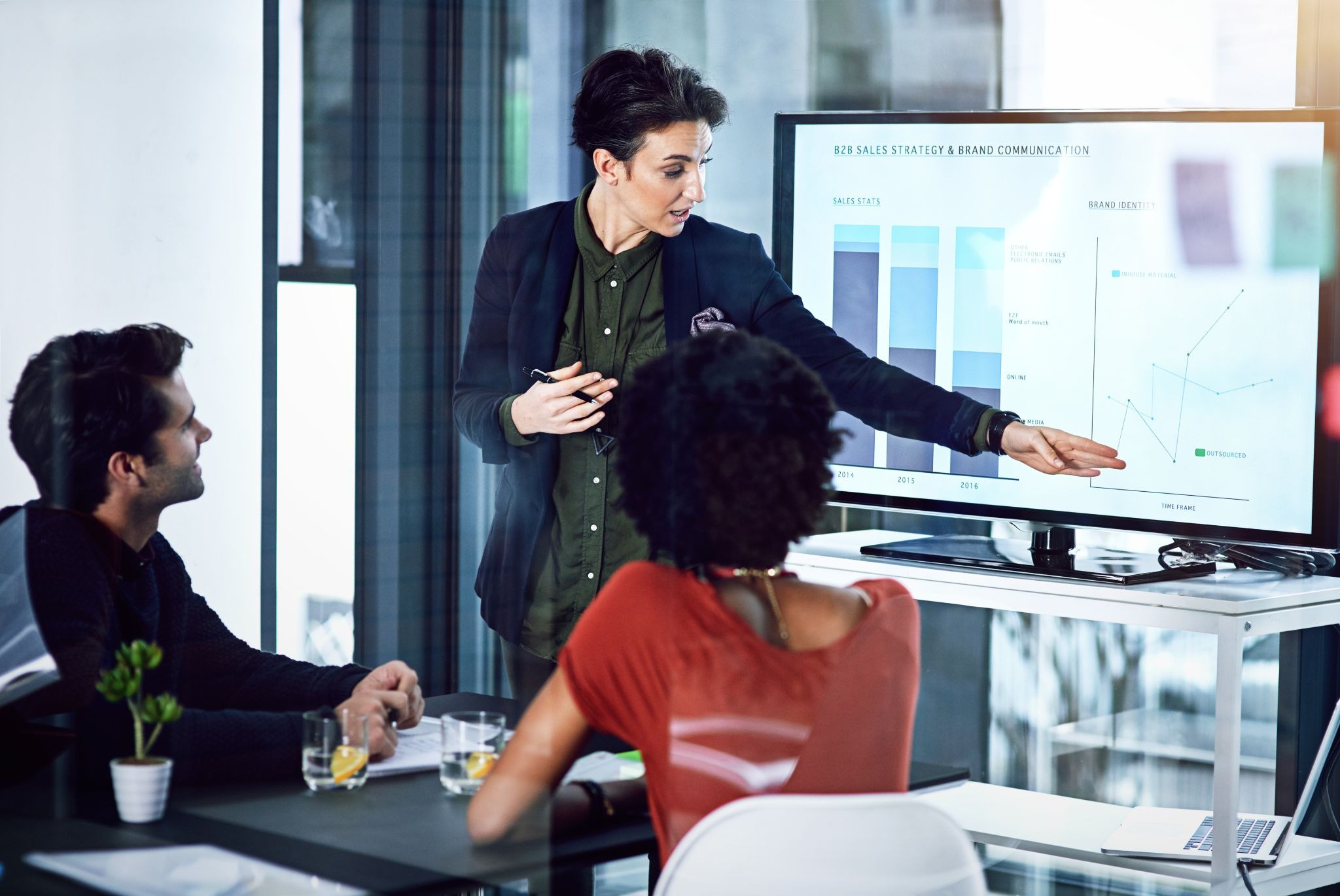 Alt text: Presenter pointing to sales and performance charts on a display while colleagues review the data.