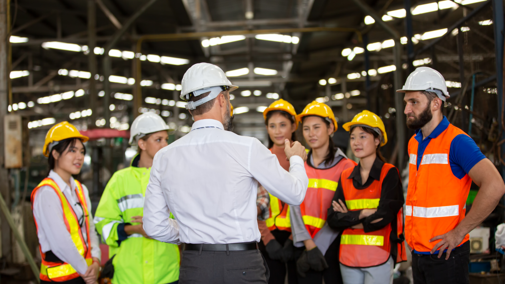 Alt text: Manager briefing a group of factory workers in hard hats and safety vests on the plant floor.