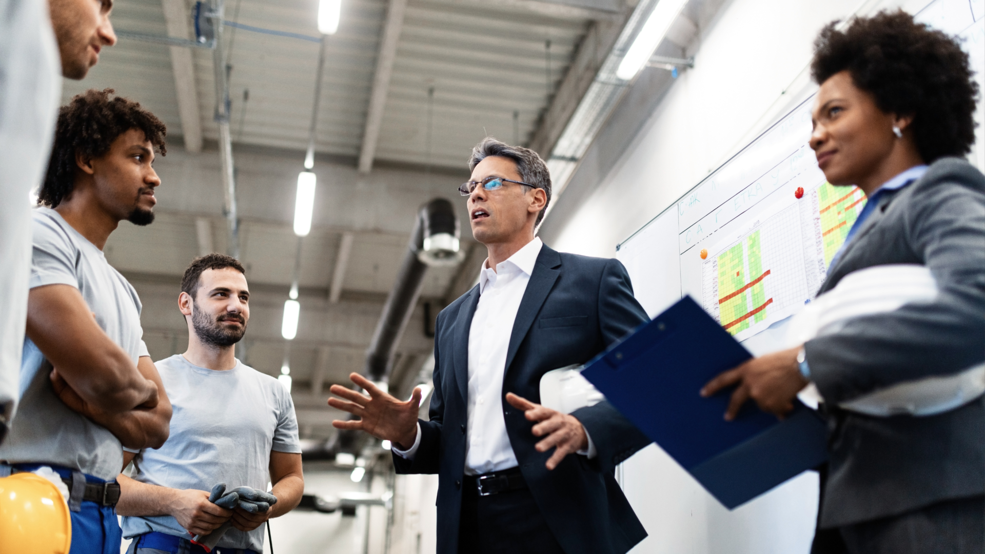 Alt text: Manager discussing a production schedule with workers in front of a planning board.