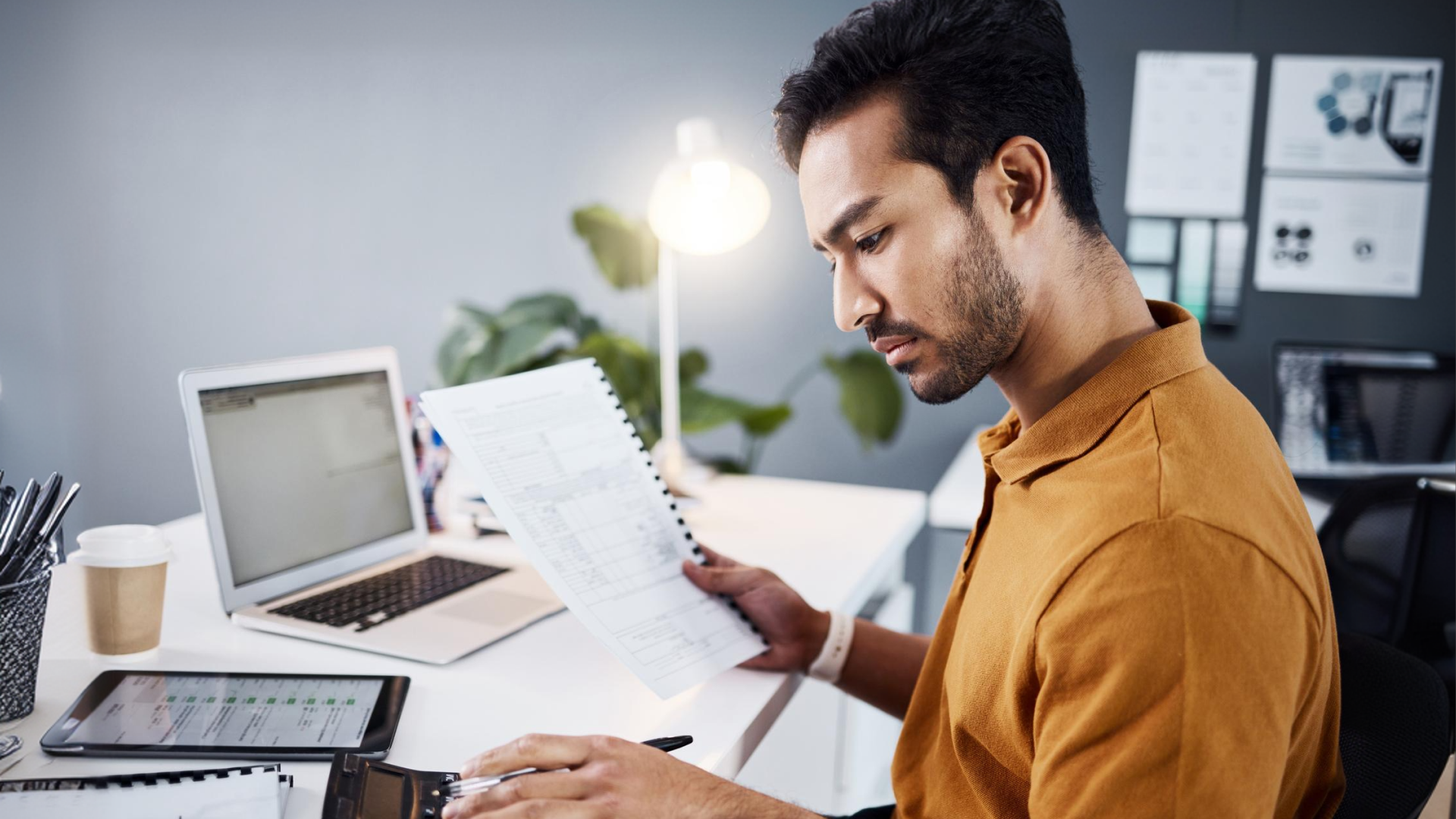 Manager reviewing a printed schedule report at his desk, with a laptop and tablet nearby.