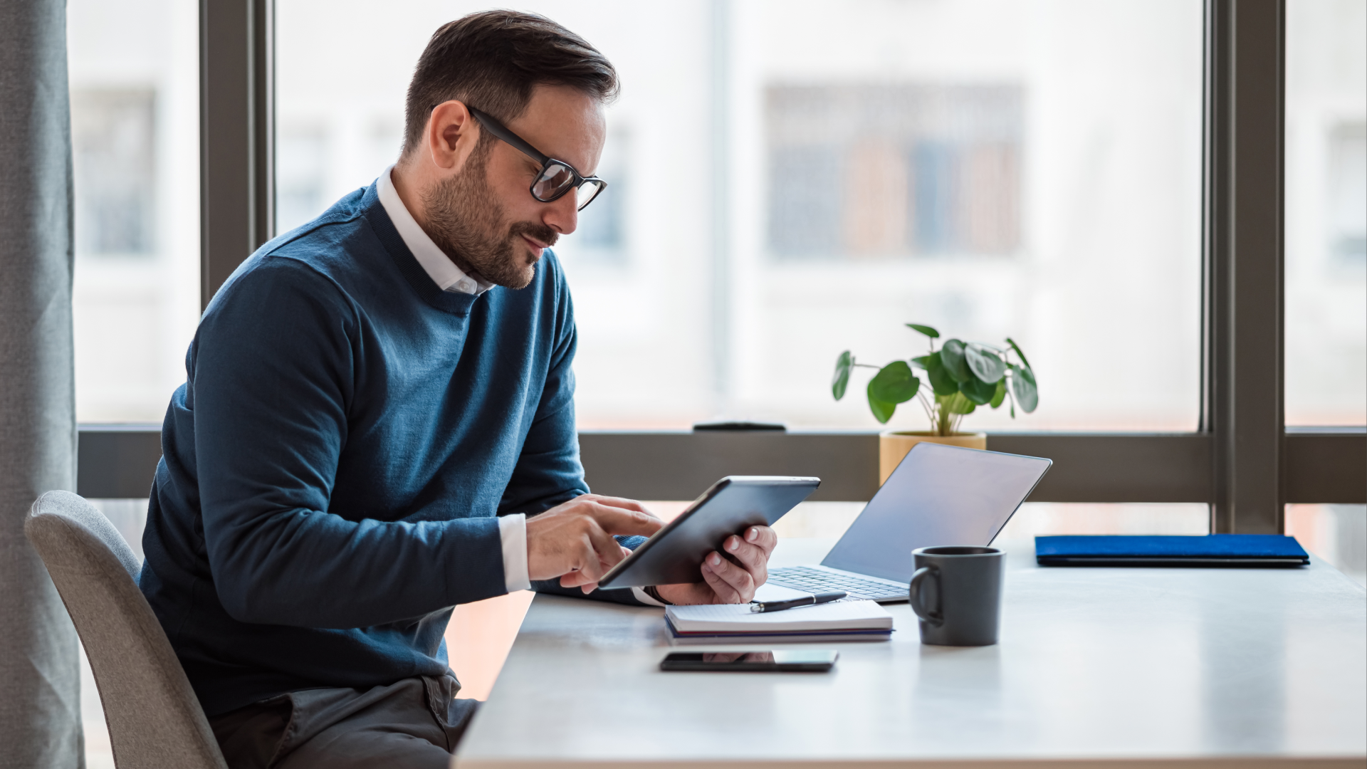 Man reviewing scheduling options on a tablet at his desk, laptop and coffee close by.