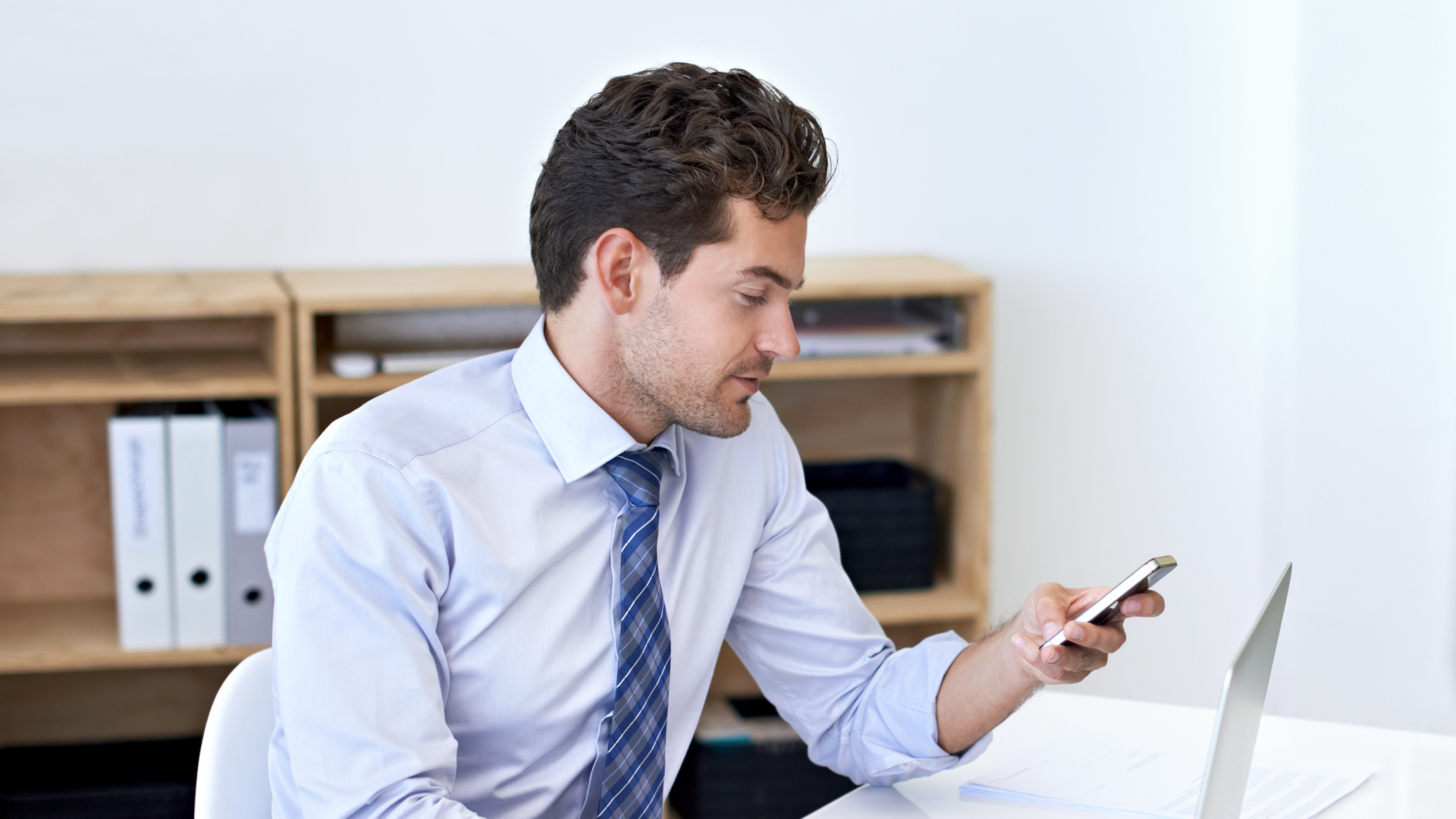 Manager checking his phone next to an open laptop while comparing scheduling apps.