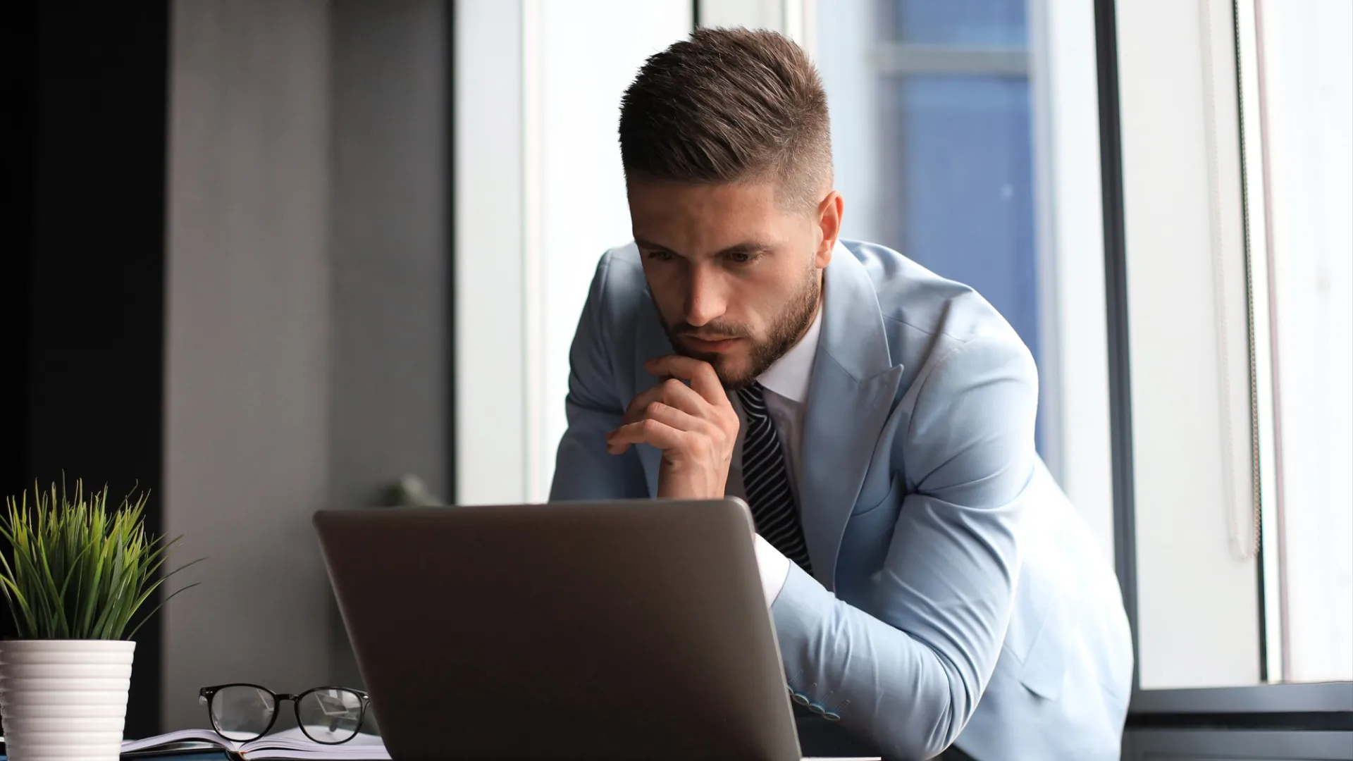 A businessman in a blue suit thoughtfully compares workforce management vs workforce optimization while looking at his laptop.