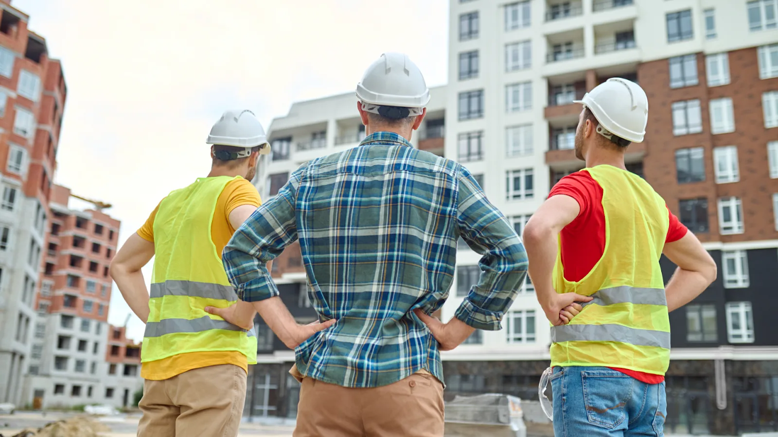 Three construction workers in hard hats and safety vests look up at a multi-story building under development.