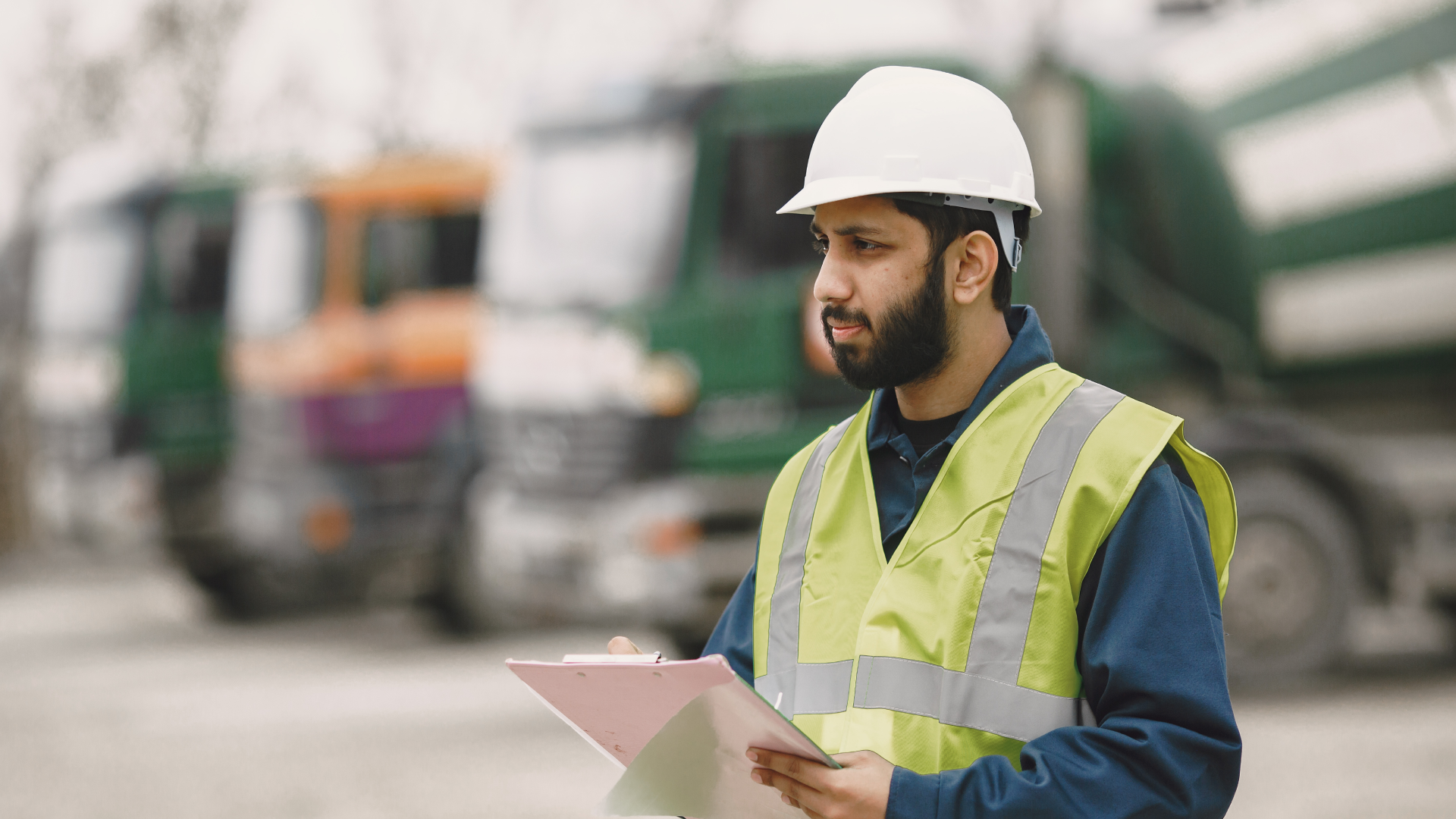 Construction worker in a hard hat and safety vest taking notes on a clipboard near job site trucks.