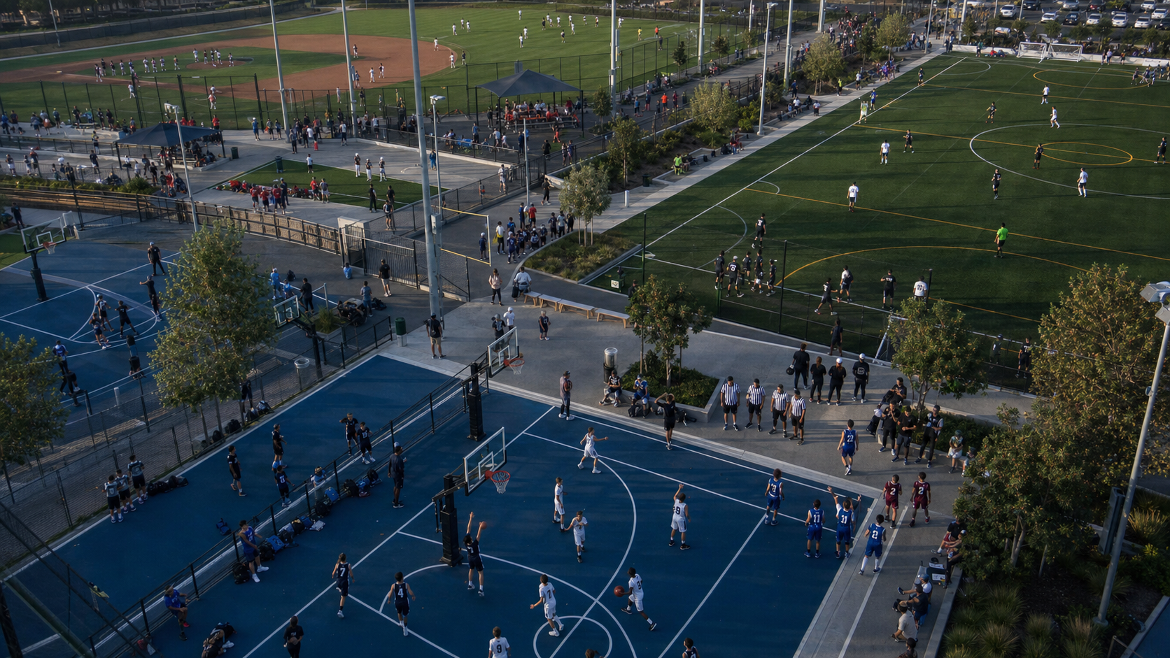 A bird's-eye view of a busy youth sports complex with multiple courts and fields visible, showing overlapping scheduling activity — coaches with clipboards, referees gathering, teams warming up in different colored jerseys, facility staff coordinating, all happening simultaneously across a large multi-venue sports facility, conveying the complexity of managing many moving parts at once, vibrant daytime lighting, dynamic composition