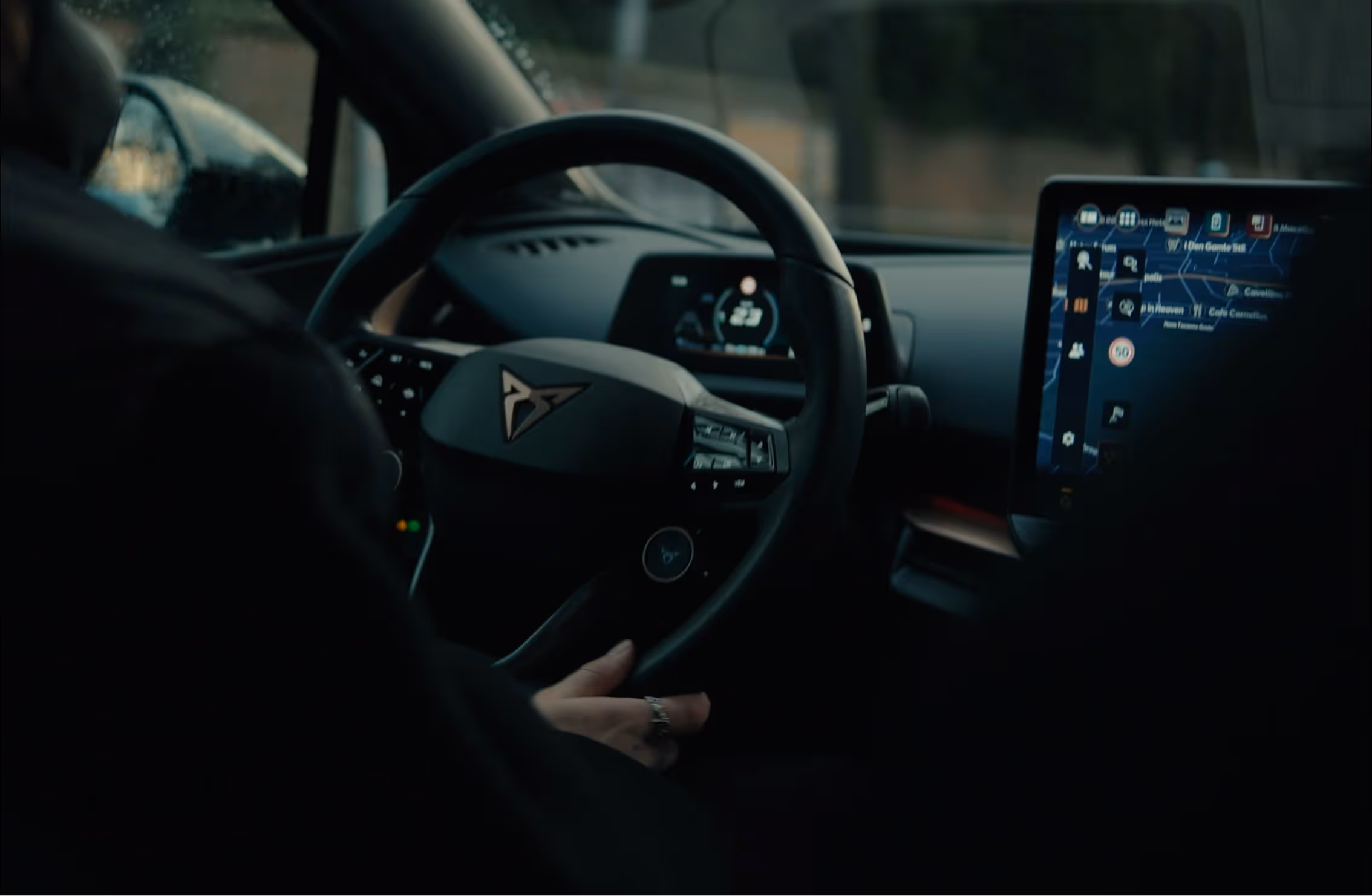 Interior view of a car showing a driver holding a steering wheel with a Cupra logo and a digital dashboard and infotainment screen displaying a navigation map.