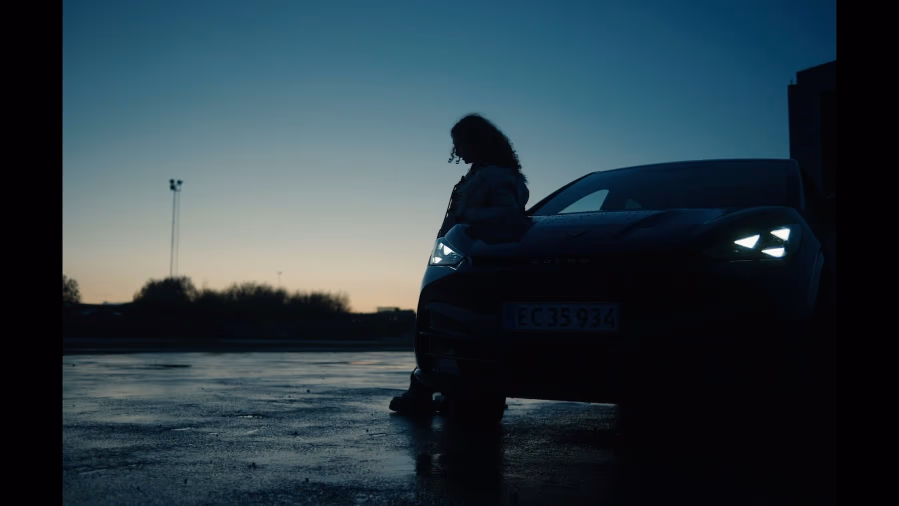 Silhouette of a person leaning against a car with headlights on at dusk in an open parking area.