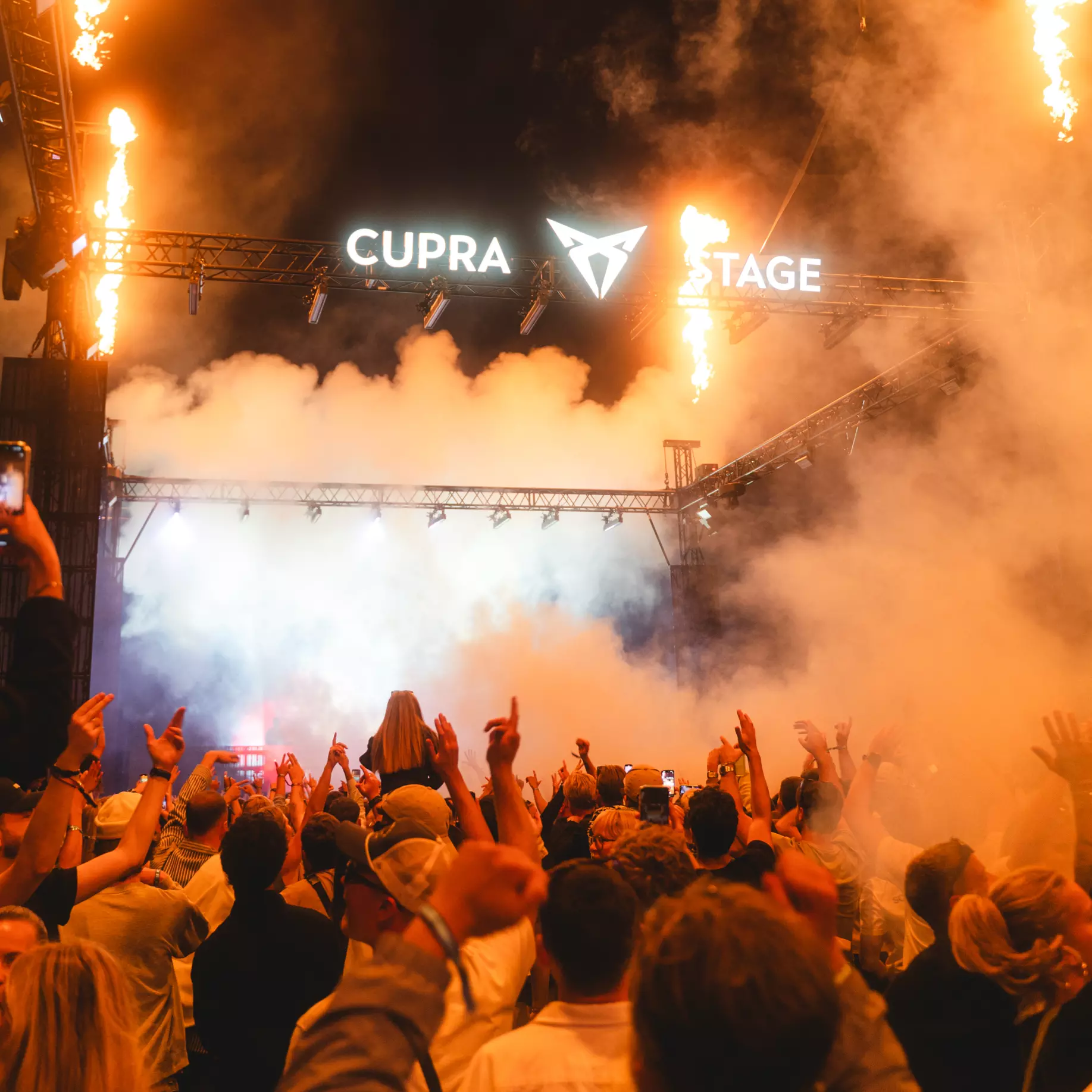 Crowd cheering with raised hands at a concert stage lit with bright lights, smoke, and flames, sign reads CUPRA STAGE.