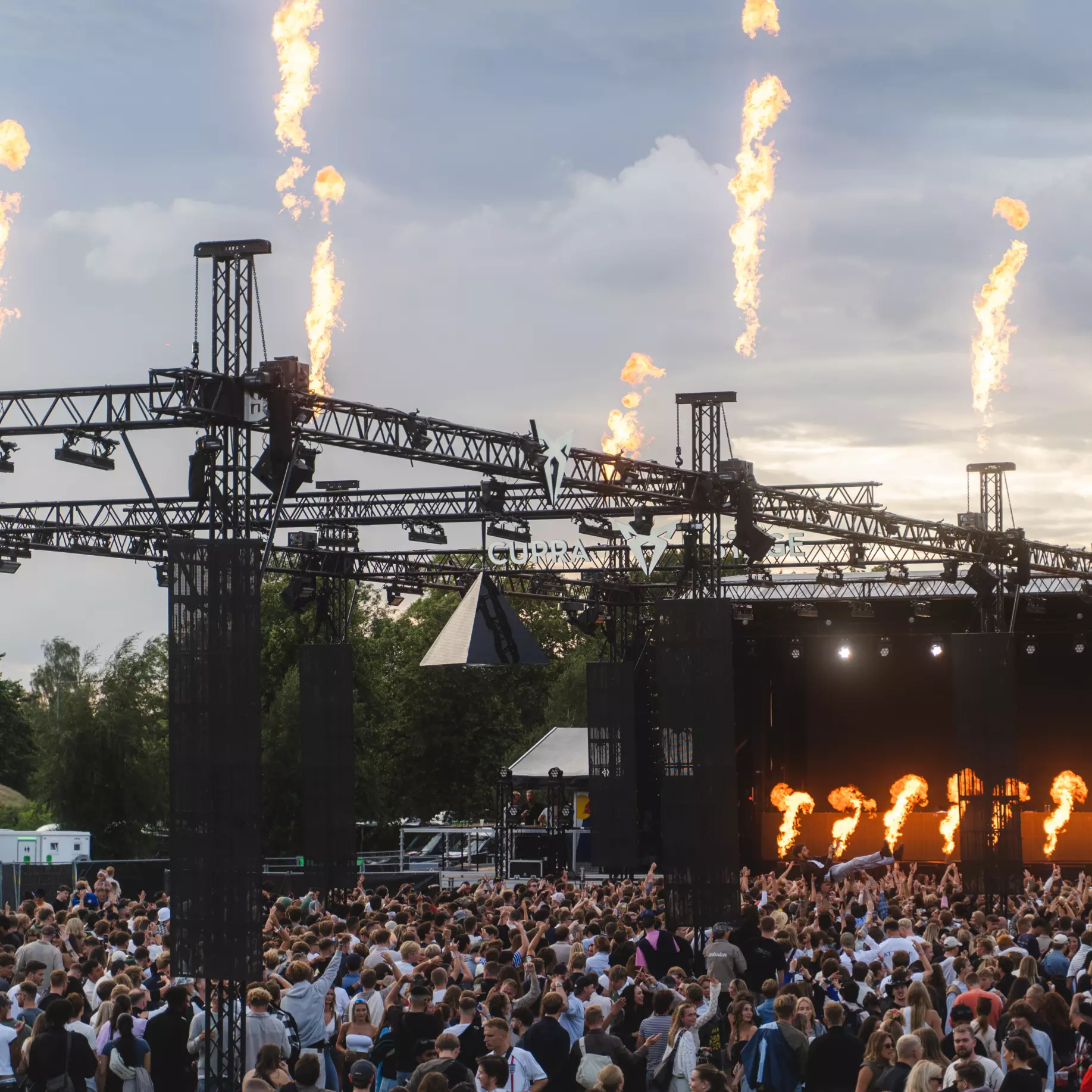 Large crowd at an outdoor music festival stage with flames shooting up from the stage lighting trusses.