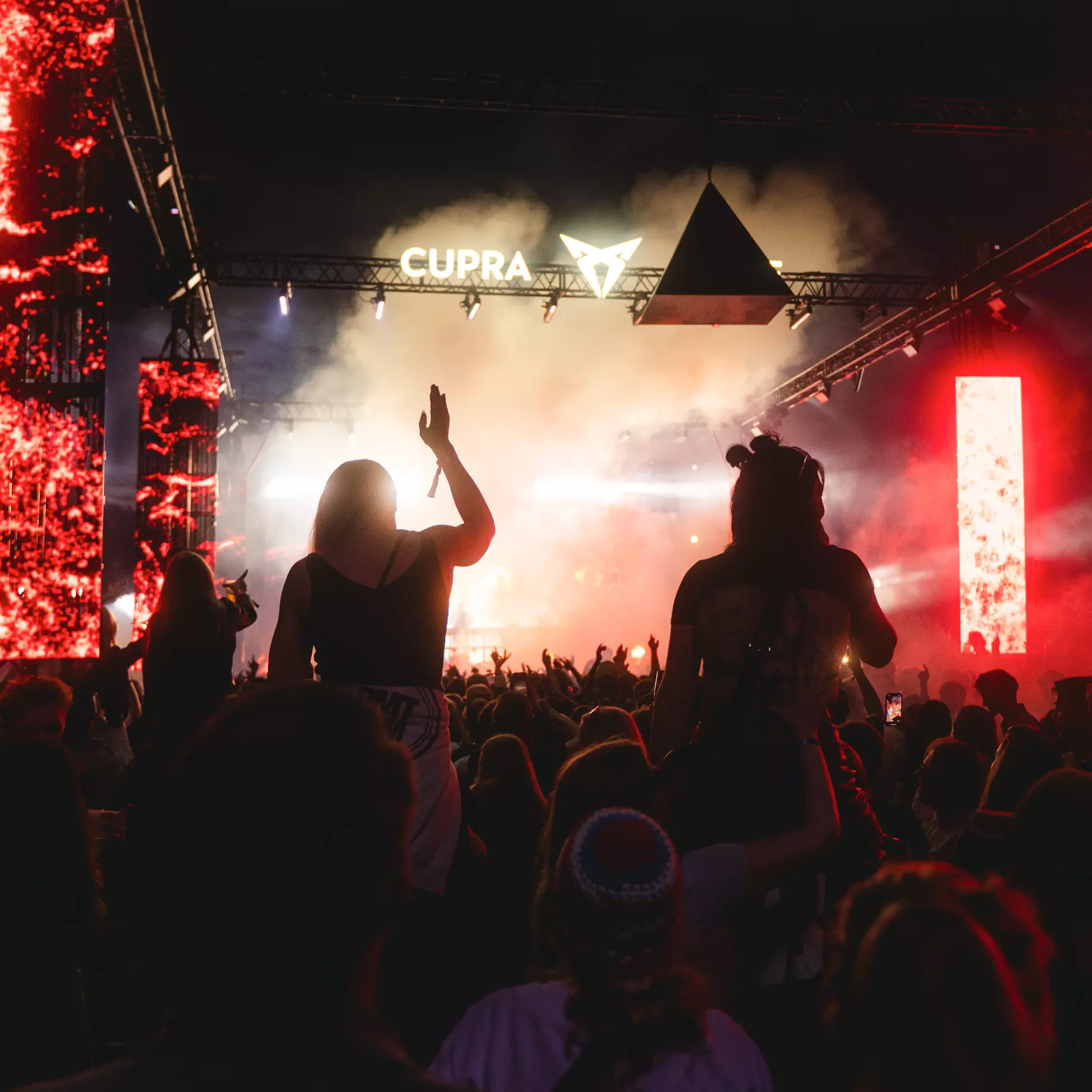 Crowd at a nighttime concert with bright stage lights, fog, and red illuminated screens.