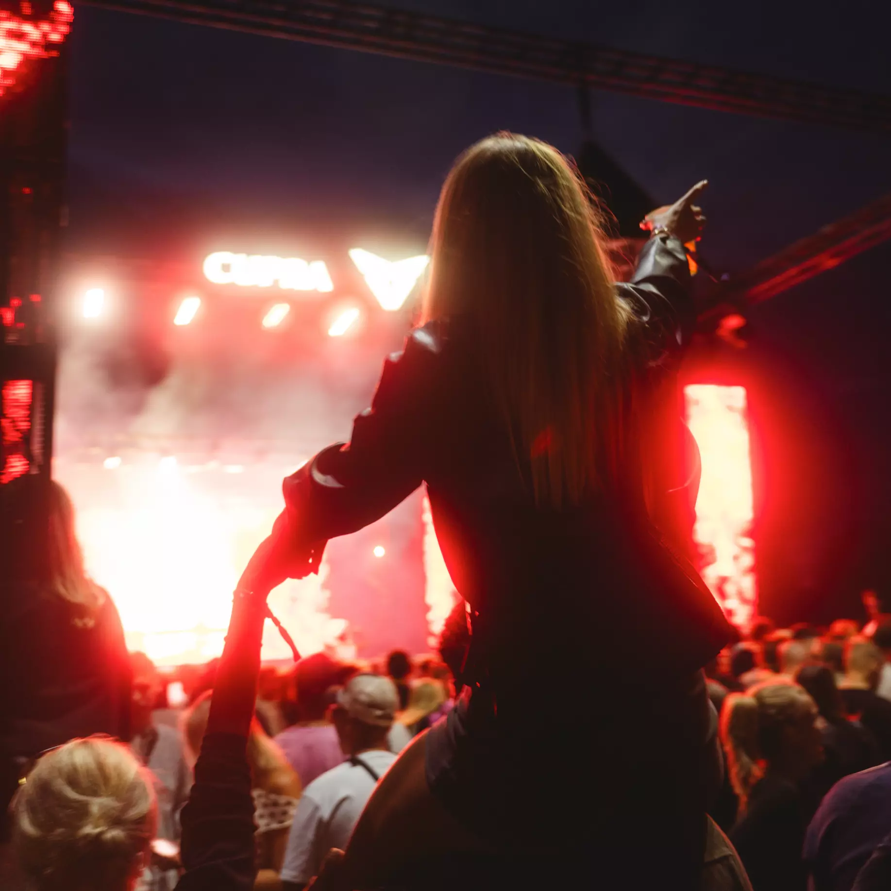 Woman on shoulders pointing at brightly lit stage during nighttime concert crowd.