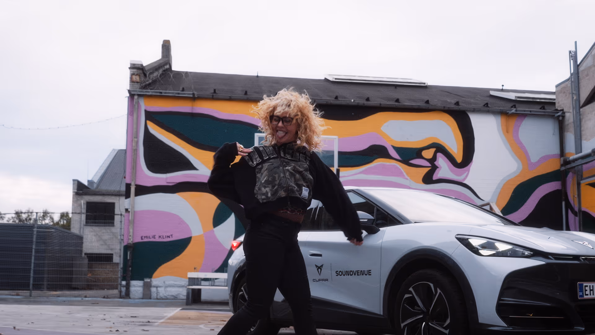 Person with curly blonde hair and glasses dancing in front of a white cupra car and a colorful abstract mural.