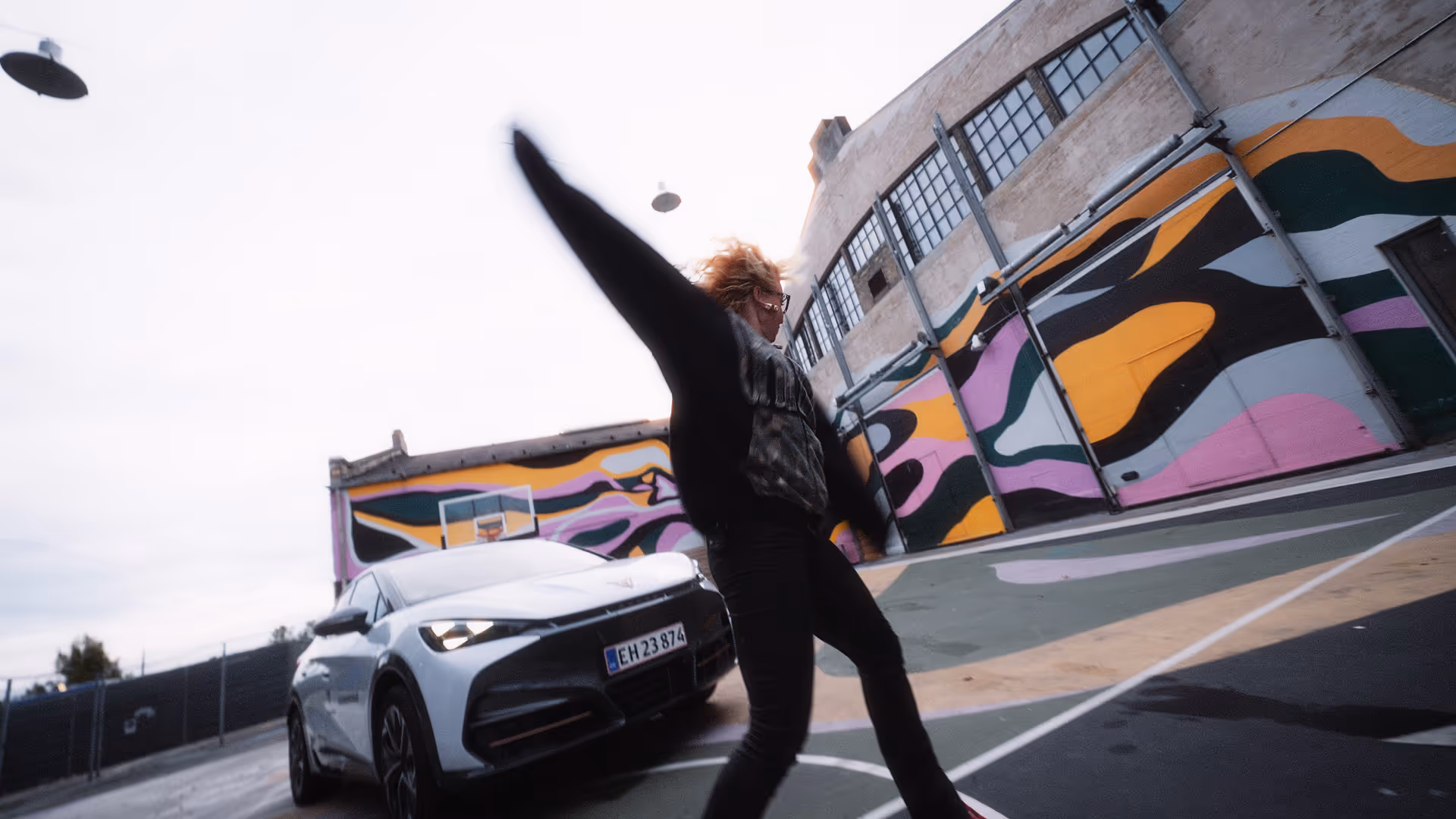 Person with outstretched arms dancing or moving dynamically on an outdoor basketball court next to a white SUV and colorful mural on a building wall.