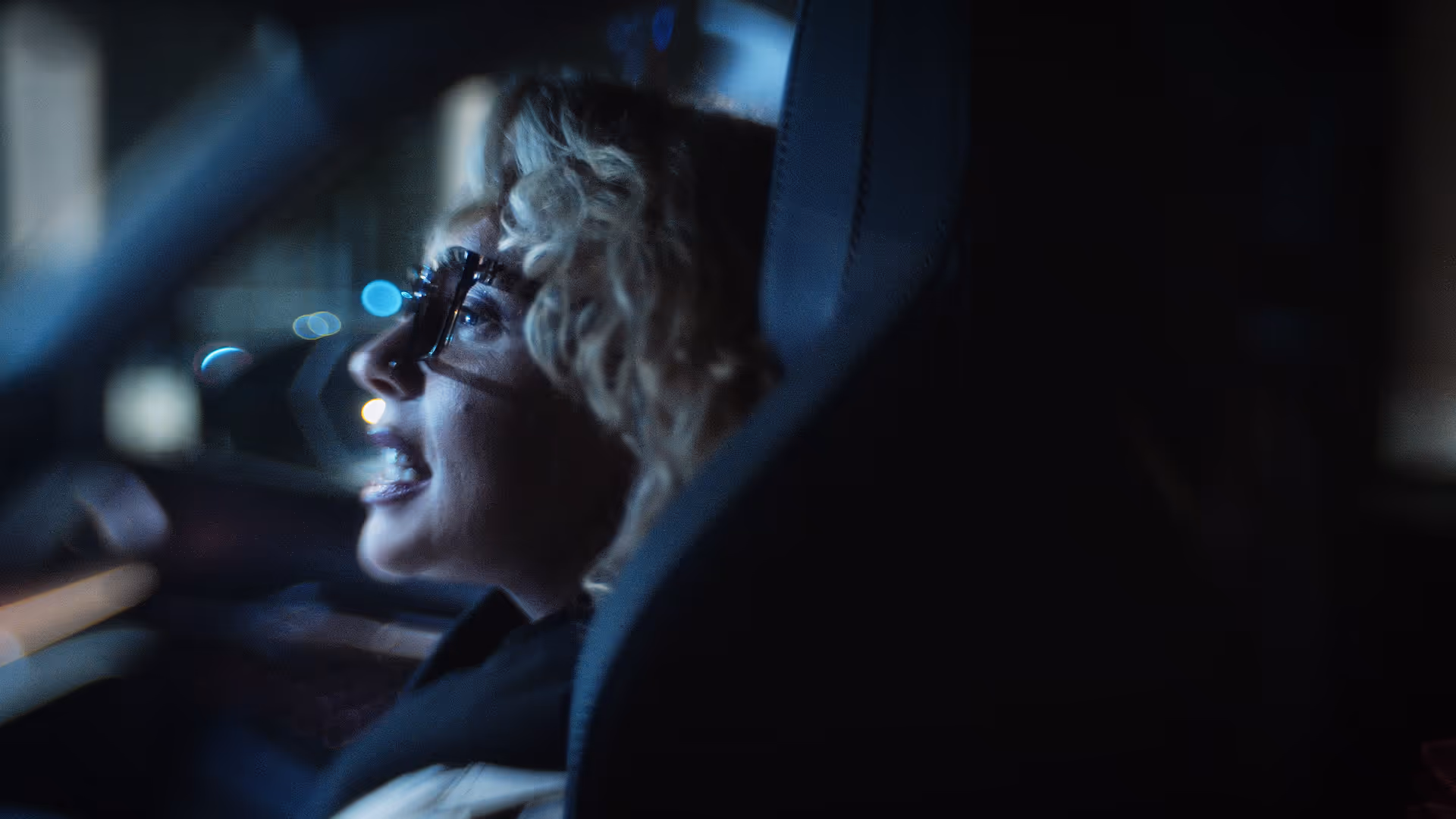 Profile of a woman with curly blonde hair and glasses smiling inside a dimly lit car at night.