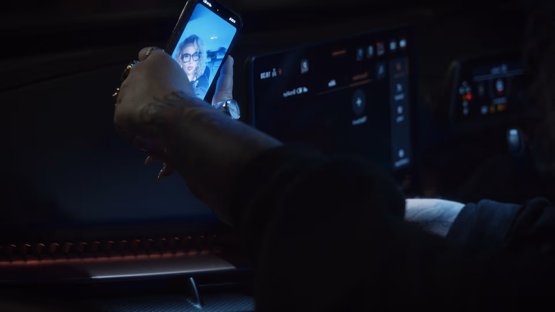 Person holding a smartphone displaying a woman with glasses on a video call inside a dark car.