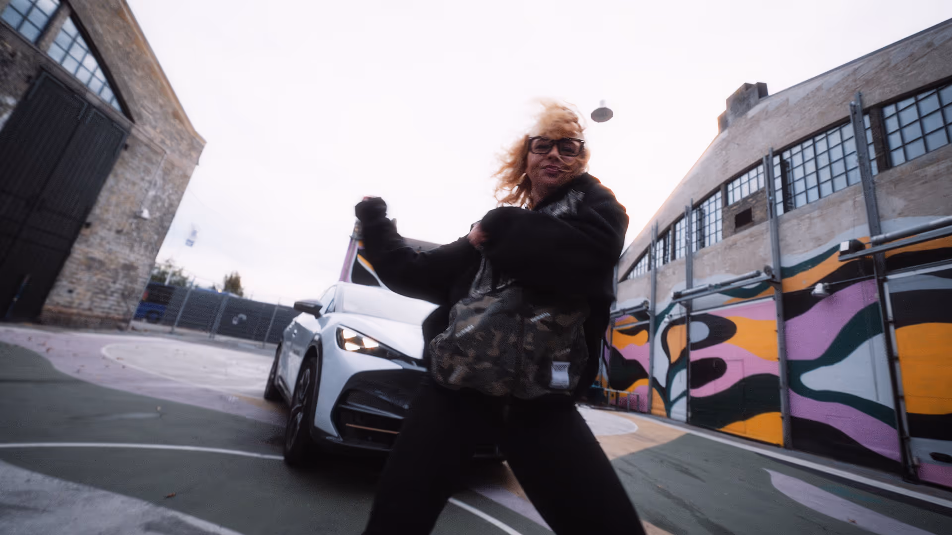 Person with curly blonde hair and glasses dancing energetically in front of a white car on a colorful outdoor basketball court.