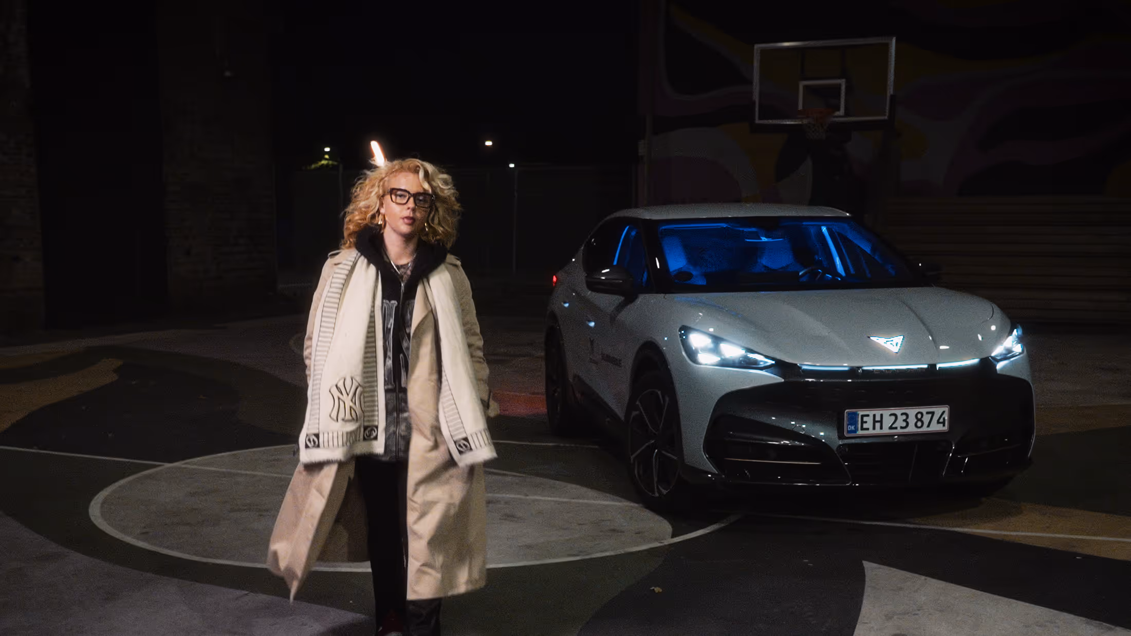 Person with curly blonde hair, glasses, beige trench coat, and white scarf walking in front of a gray modern car with headlights on at night on a basketball court.