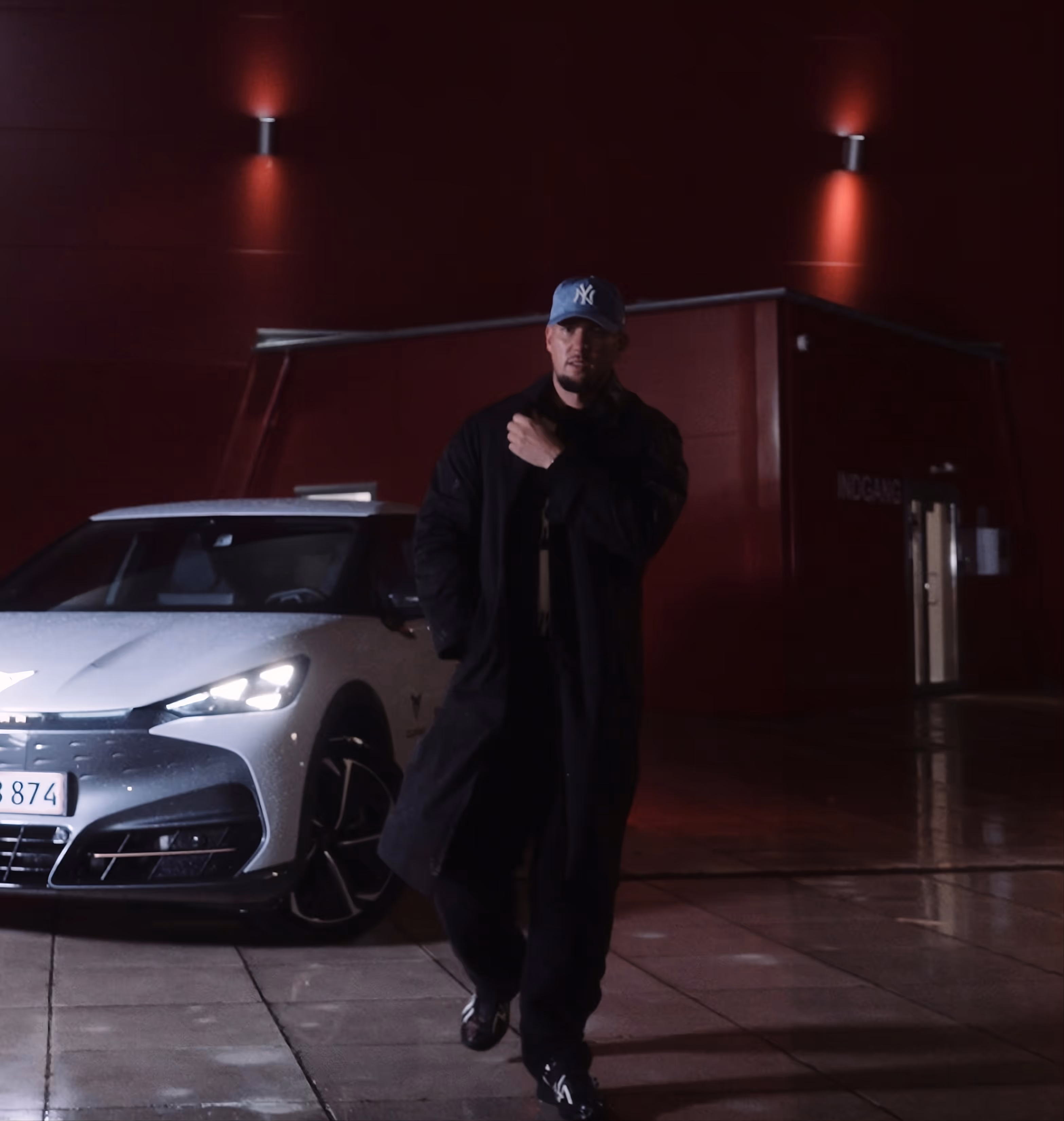 Man in a black coat and blue cap walking on wet pavement near a white car at night.