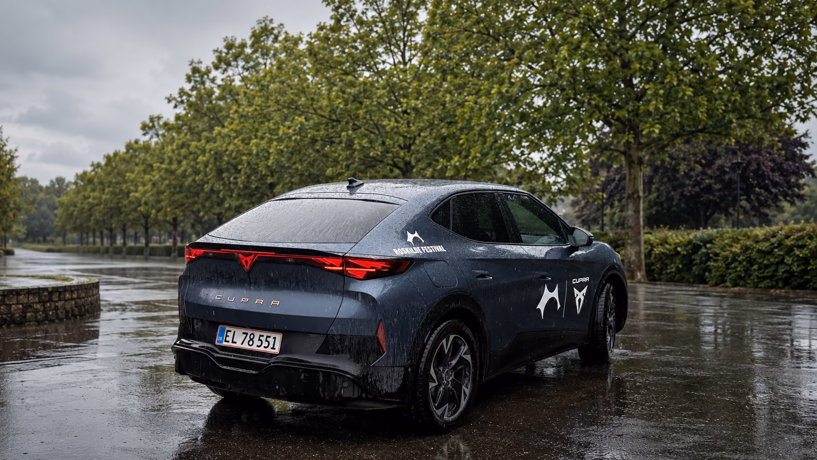 Rear-side view of a blue Cupra electric car parked on a wet road with trees in the background.