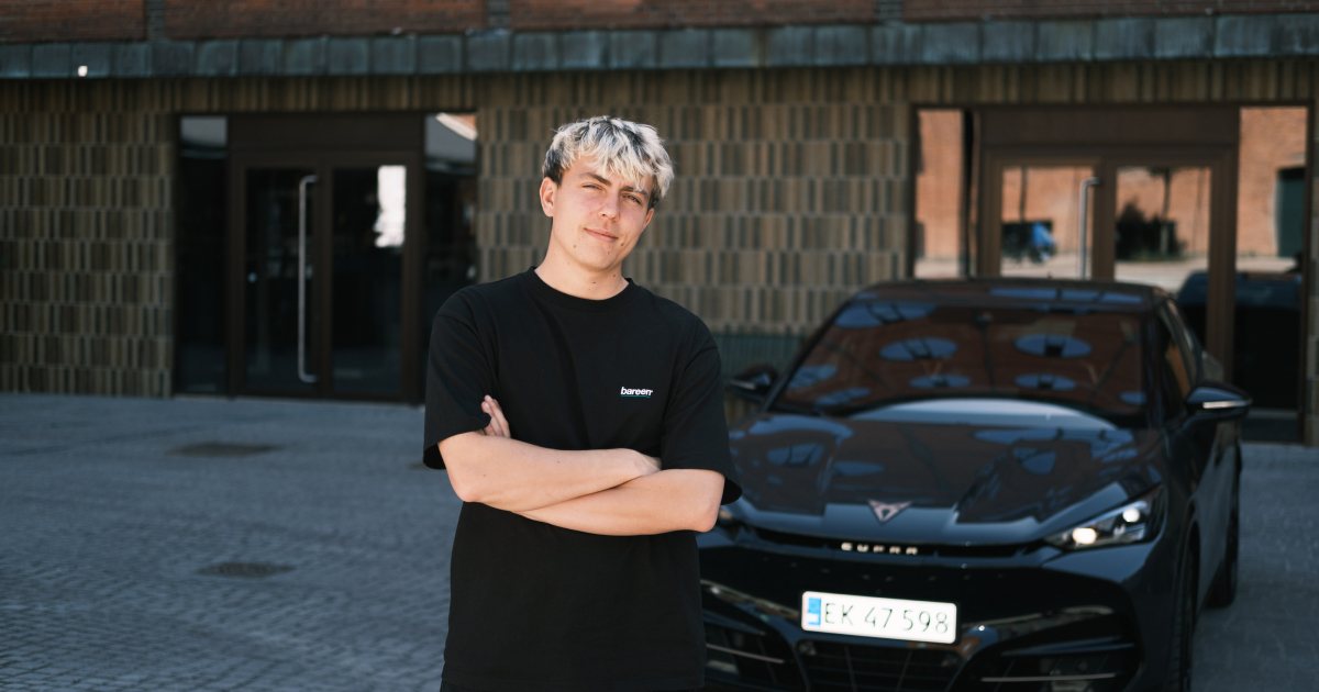 Ung mand med blondt hår står med armene krydsede foran en sort bil i en gårdsplads.