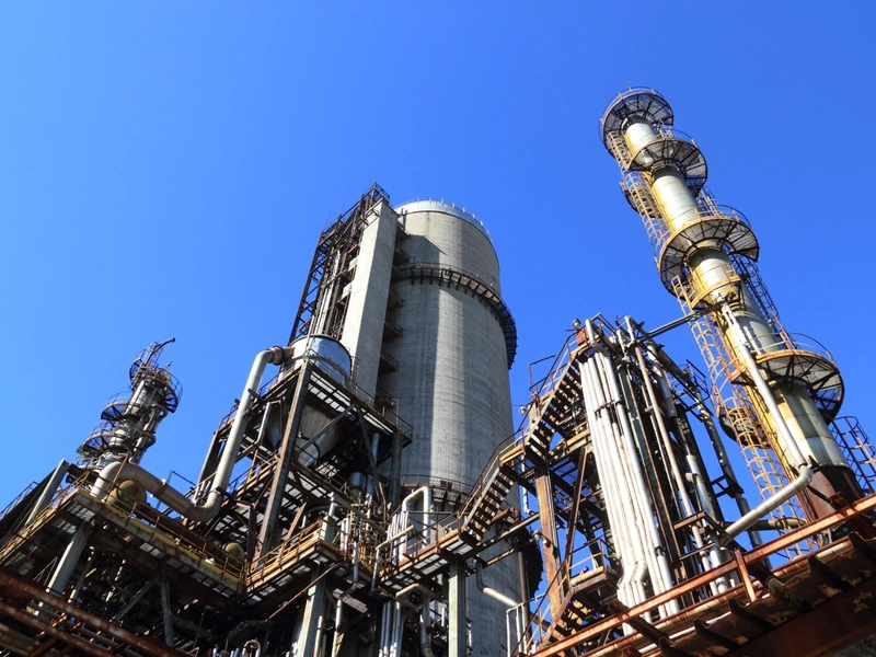 Planta industrial con altas estructuras metálicas y tuberías contra un cielo azul claro.