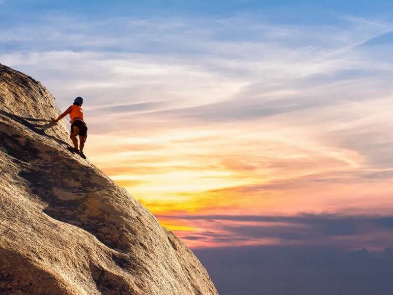 Una persona con equipo de escalada asciende por una empinada pared rocosa contra un vibrante cielo al atardecer.
