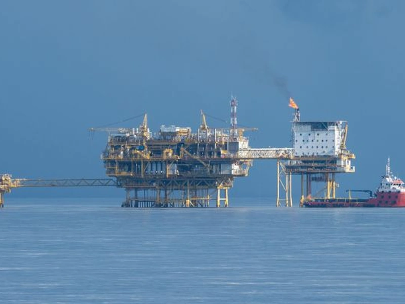Plataforma petrolífera en alta mar con una pila de bengalas que quema gas, junto a un buque de suministro rojo en un mar tranquilo.