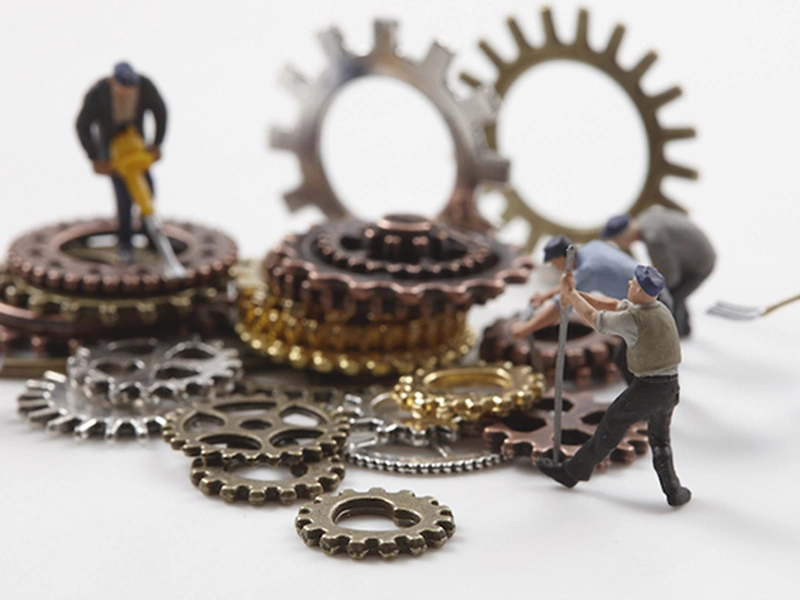 Figuras en miniatura trabajando entre engranajes metálicos de varios tamaños.
