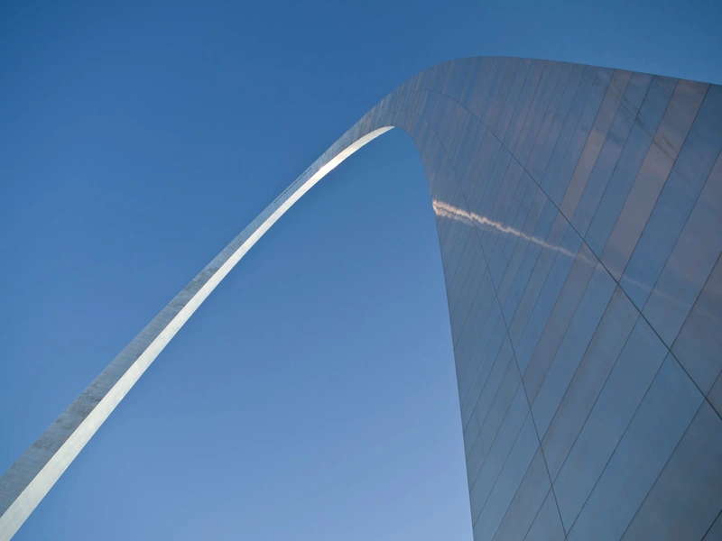 Vista de un gran arco de acero contra un cielo azul claro, con una tenue estela que atraviesa la estructura.