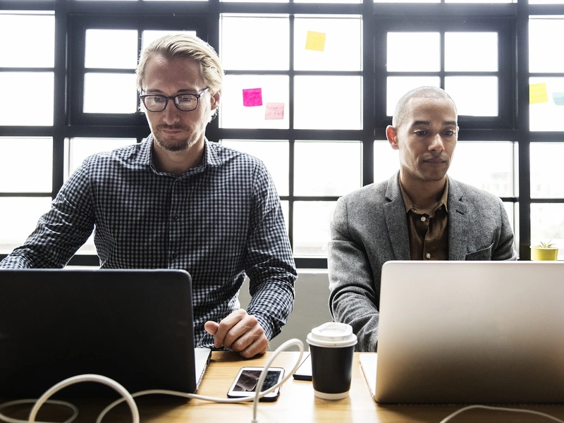 Dos hombres sentados en una mesa, cada uno trabajando en una computadora portátil.