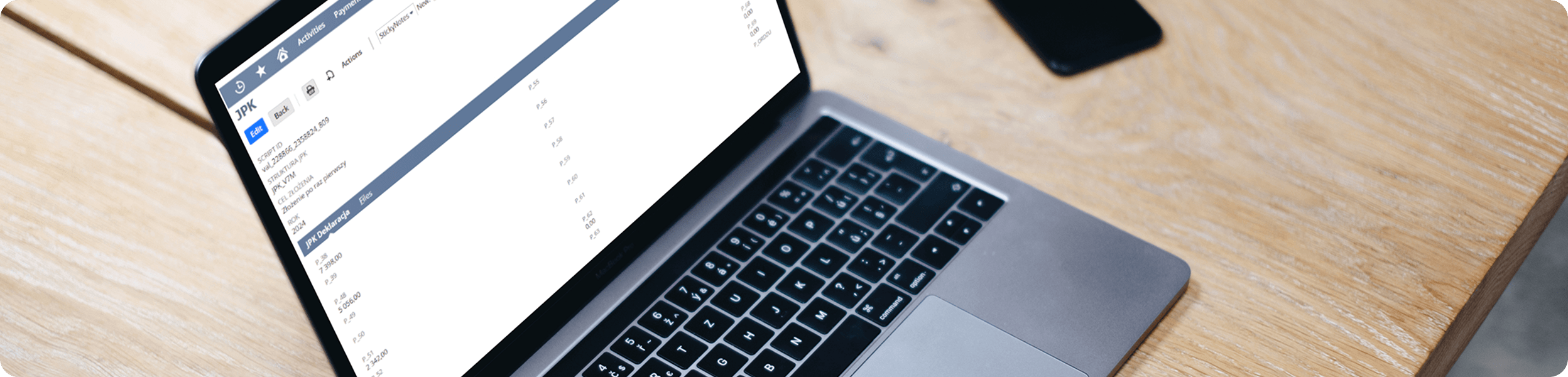 Młoda kobieta z blond włosami i okularami czyta dokument przy biurku z komputerem.