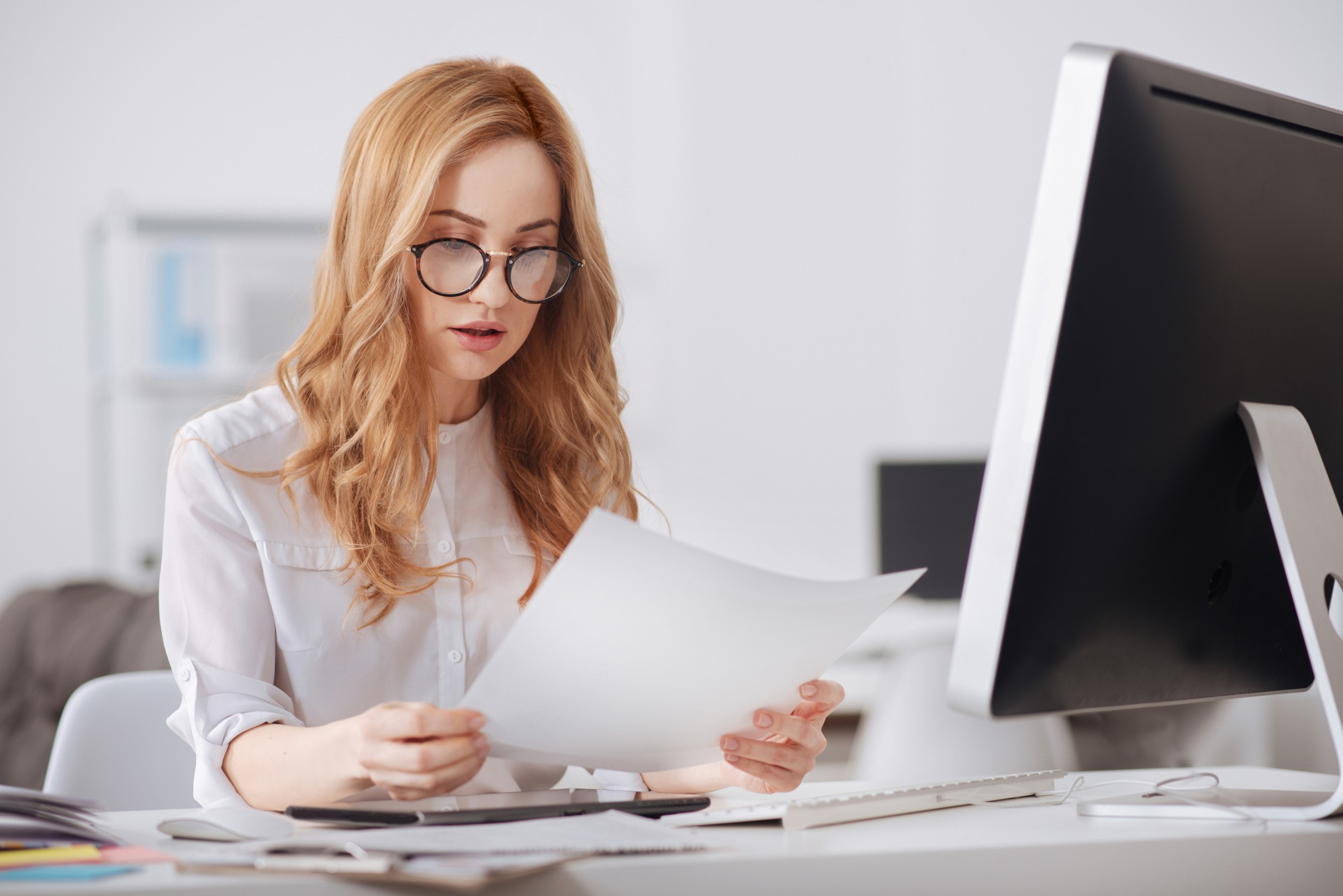 Młoda kobieta z blond włosami i okularami czyta dokument przy biurku z komputerem.