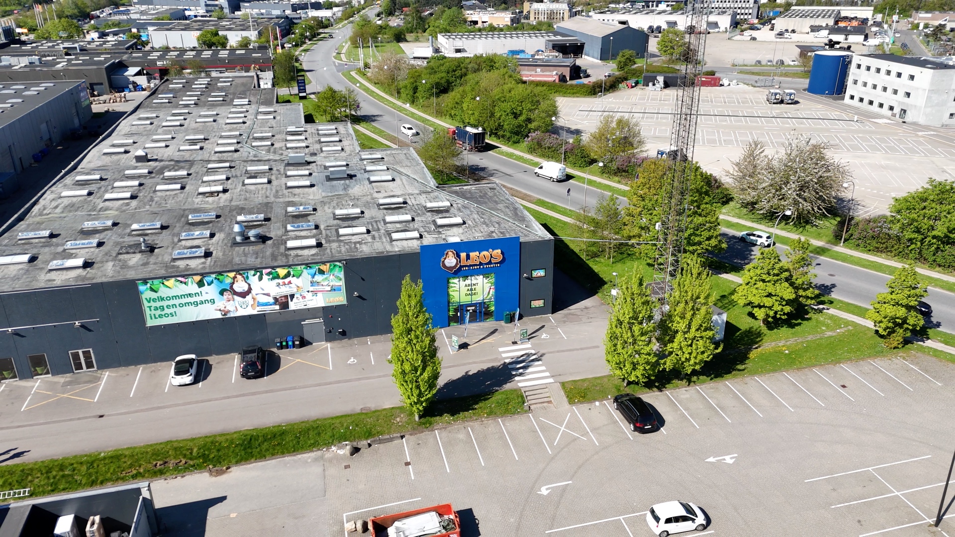 Luftfoto af Leo's legeland og eventcenter med parkeringsplads og omkringliggende veje og træer.