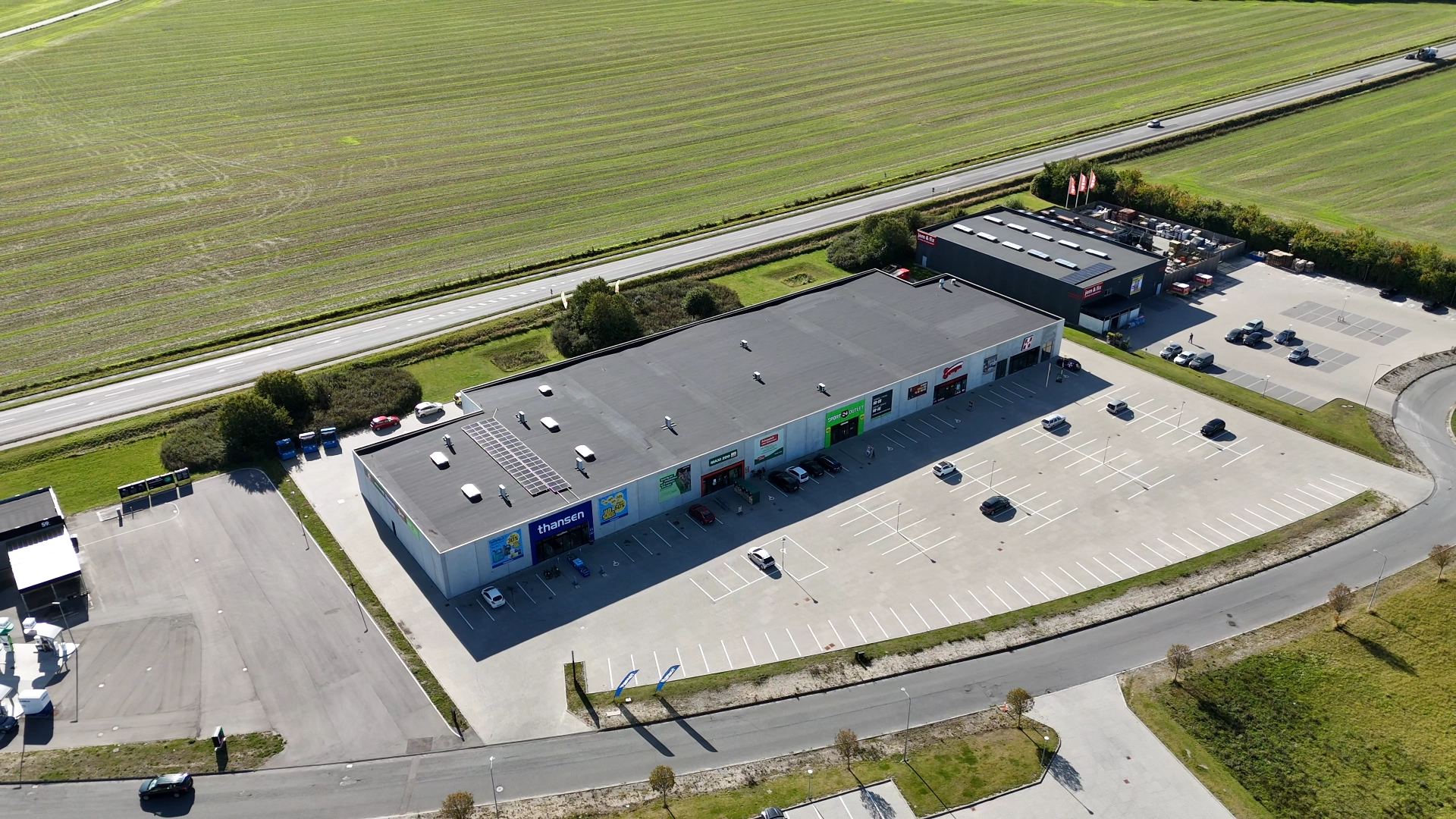Luftfoto af et kommercielt område med flere butikker, parkeringsplads med få biler og marker i baggrunden.