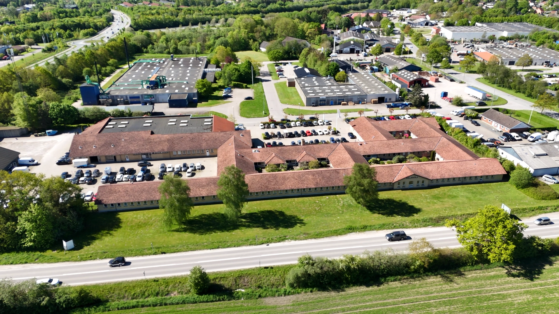 Luftfoto af kontor- og industribygninger med parkeringspladser, omgivet af grønne områder og veje.