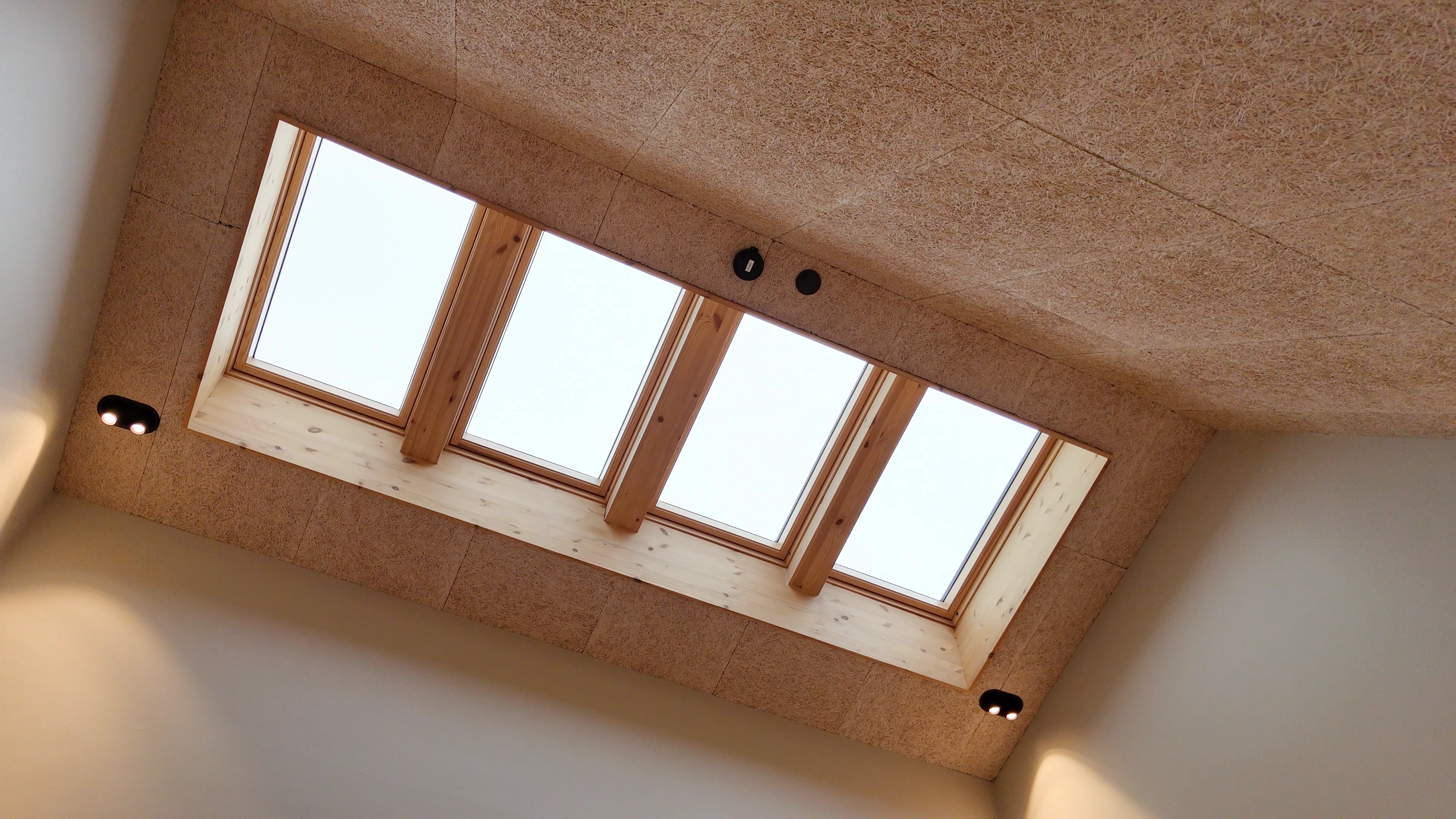 Loftvindue med fire glasruder i et træramme, monteret i et loft med lyse brune akustikpaneler og to sorte loftslamper.
