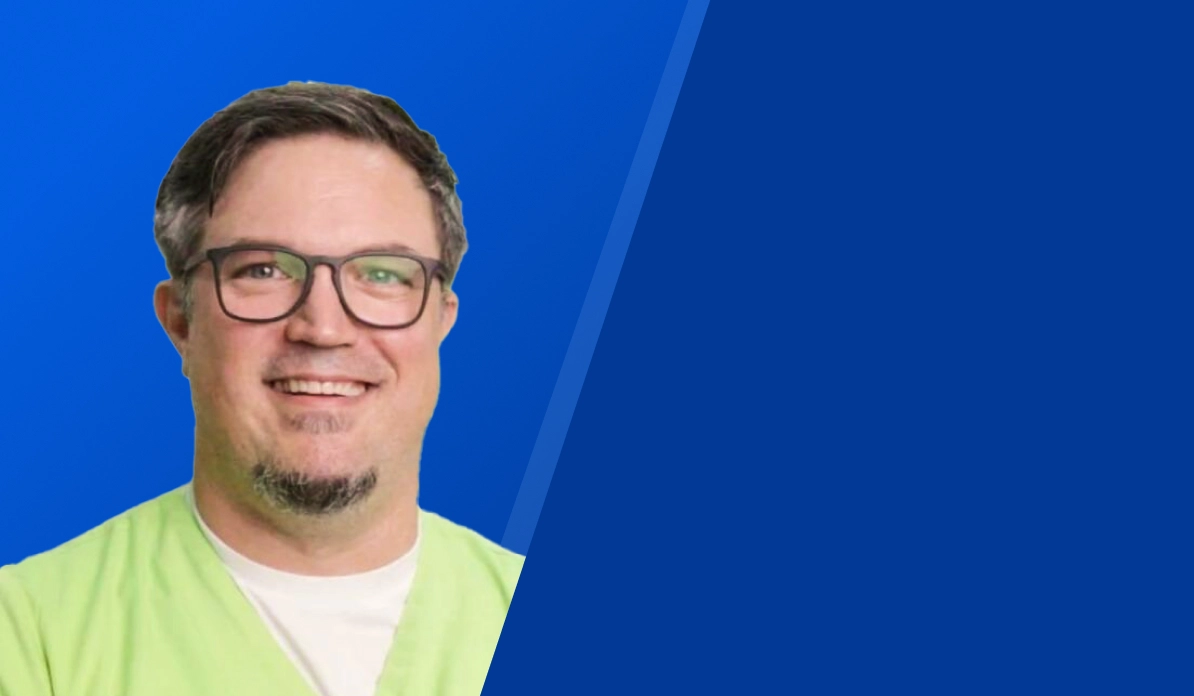 Porträt eines lächelnden Mannes mit Brille und grünem Oberteil vor blauem Hintergrund.