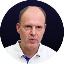 Frau mit dunklen lockigen Haaren, Brille und heller Bluse vor schwarzem Hintergrund.