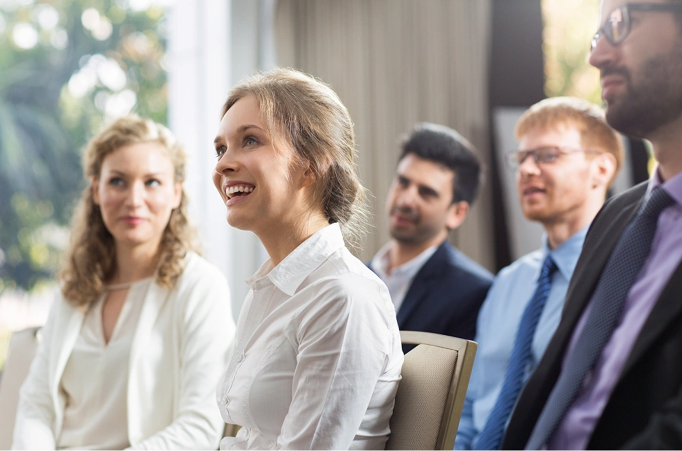 Mehrere lächelnde Erwachsene sitzen in einer Konferenz oder Besprechung in einem hellen Raum.