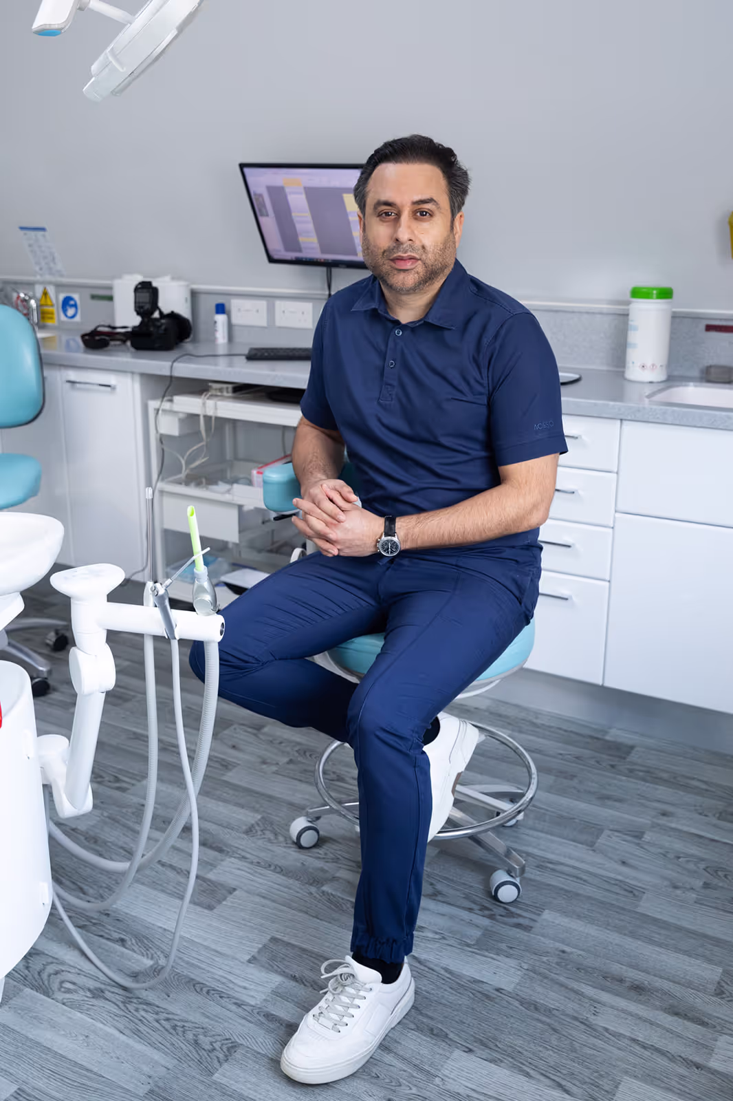 Man in navy blue scrubs sitting on a stool in a dental office with dental equipment and a computer monitor in the background.