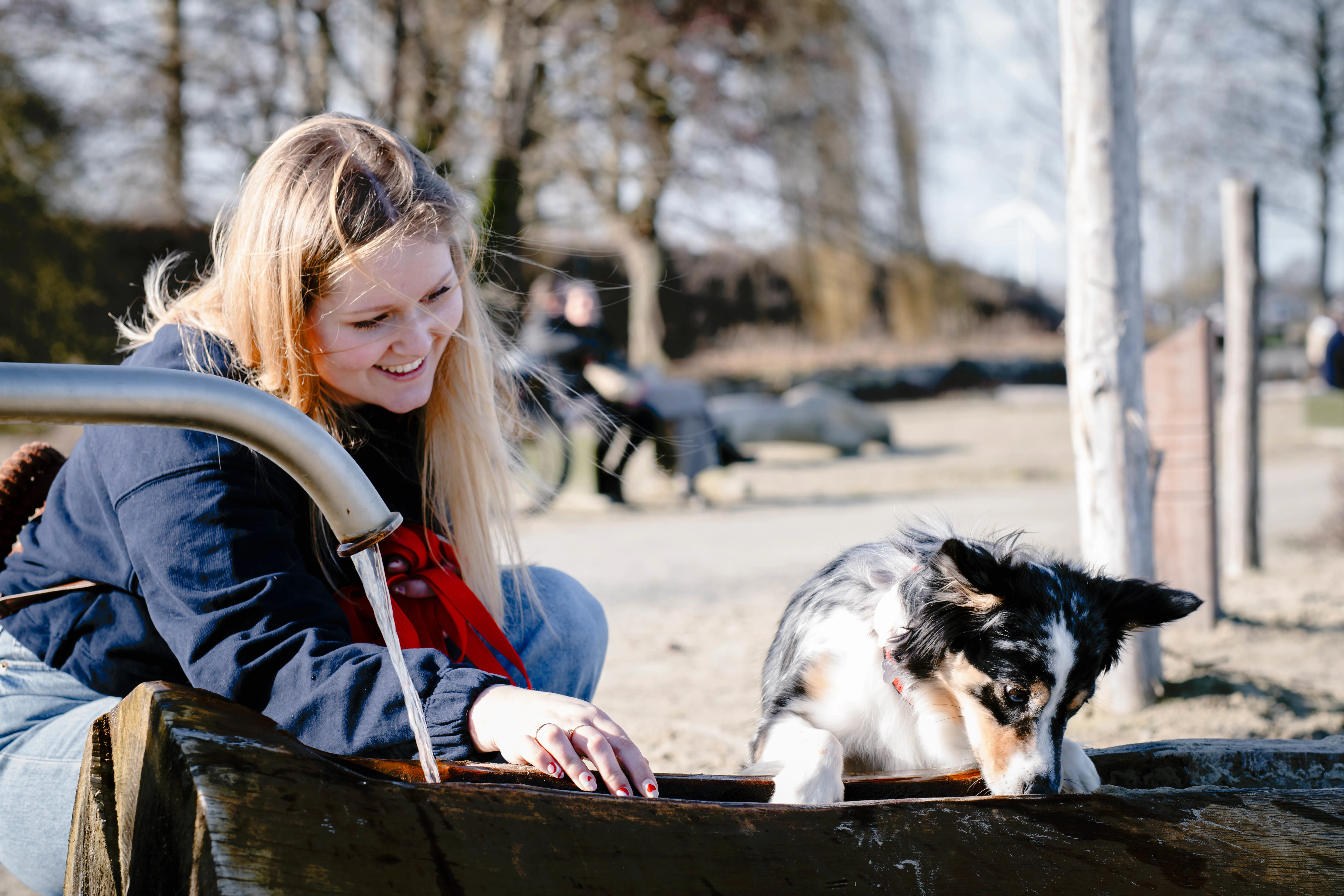Vrolijke vrouw met lang blond haar die knielt bij een houten waterbak met een stromende metalen waterkraan, terwijl een zwart-witte hond met bruine accenten uit de waterbak drinkt in een parkachtige omgeving.