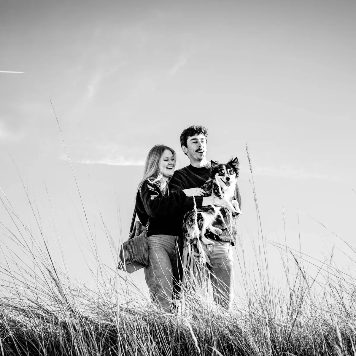 Jong koppel op een heuvel met hoog gras, waarbij de man een middelgrote hond vasthoudt terwijl ze beiden lachen onder een bewolkte hemel.