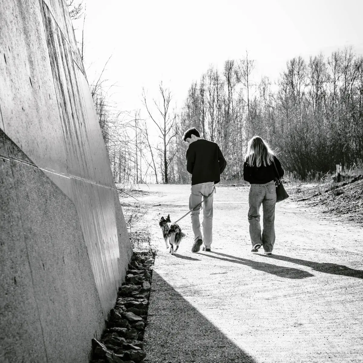 Zwart-witfoto van een jong koppel die een hond uitlaat op een bospad naast een grote betonnen muur, met kale bomen op de achtergrond.